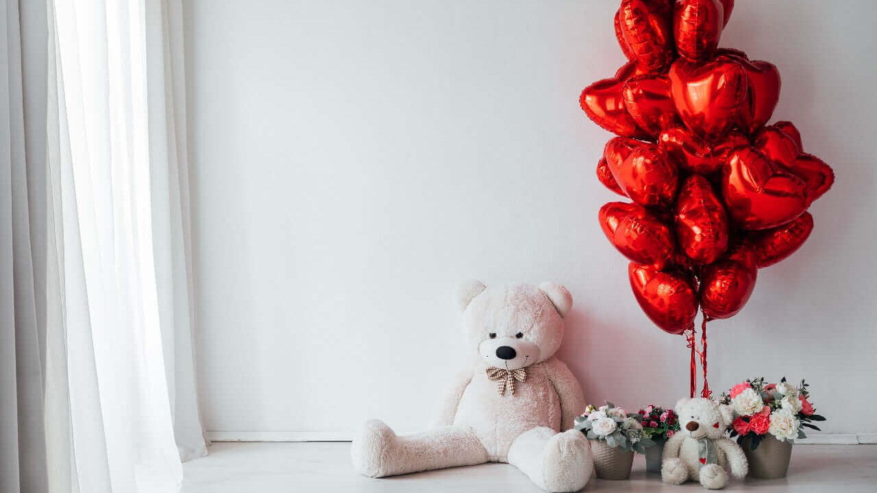 A cozy setup featuring a large white teddy bear, a bunch of shiny red heart-shaped balloons, and flower arrangements in neutral-toned pots, placed against a minimalist white background with sheer curtains.