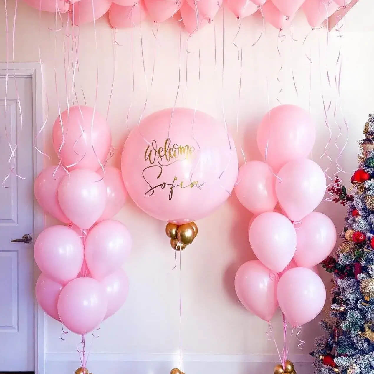 Pink balloon arch with 'Welcome Sofia' text, Christmas tree on right