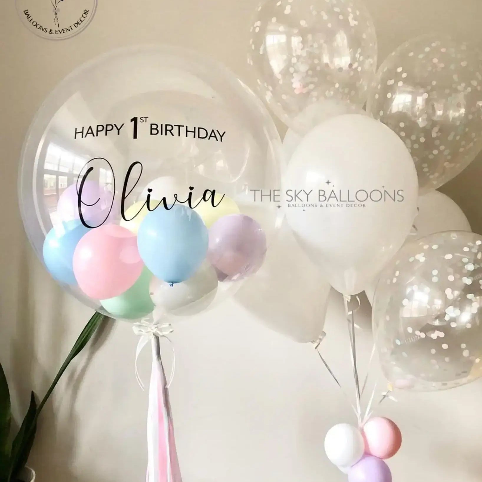 A balloon bouquet featuring a mix of round and oval-shaped balloons in shades of pink, white, blue, and green, with 'Happy 1st Birthday' and a name 'Olivia' printed on a pink banner.