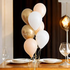 A bouquet of helium-filled balloons in various colors, including gold, white, and pink, tied together and placed on a table.