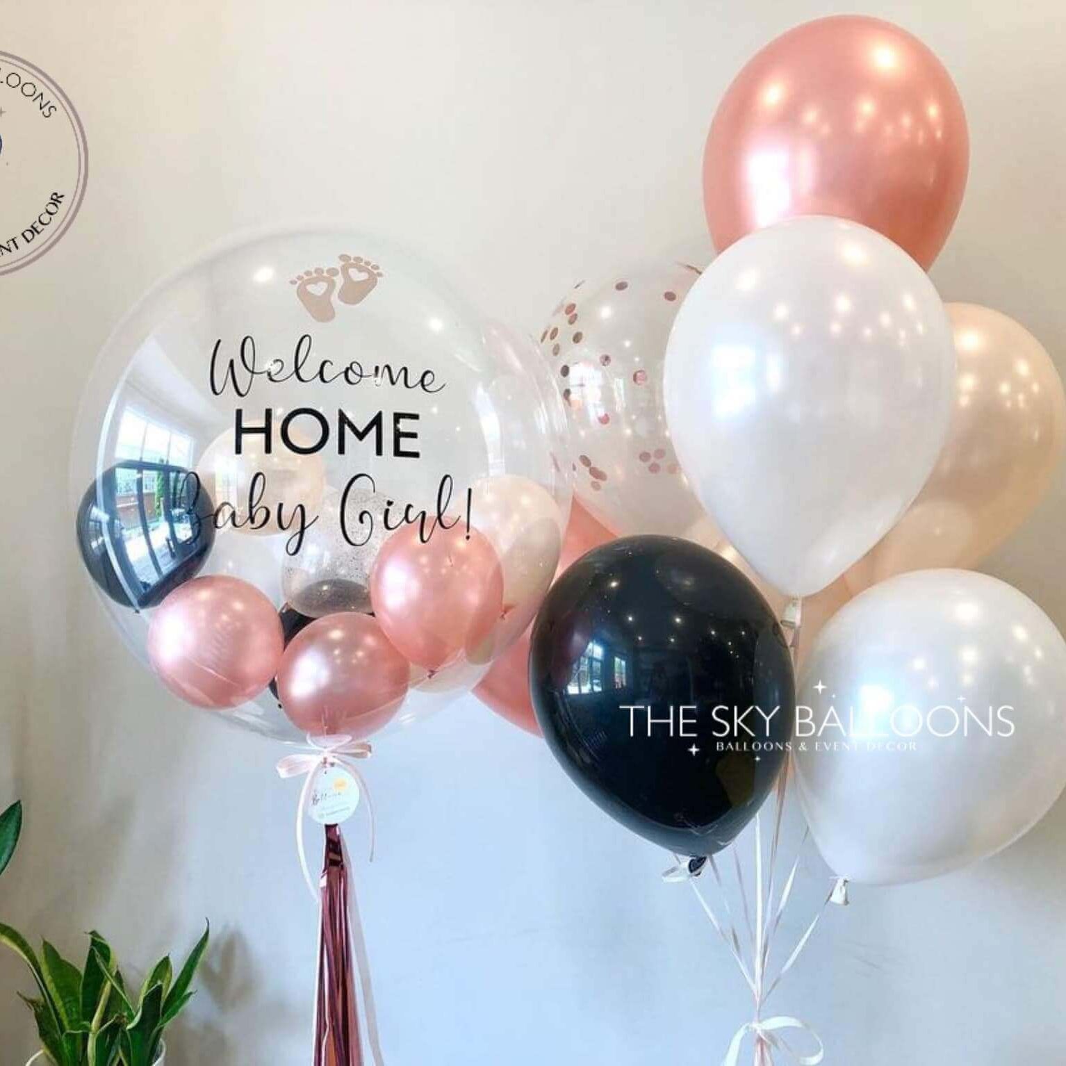A bouquet of latex balloons in various shades of pink, white, and black, filled with helium gas, with customized text that reads 'Welcome Home Baby Girl'. The balloons are arranged in a bunch with a pink ribbon and pearls, and the brand 'The Sky Balloons' is visible.