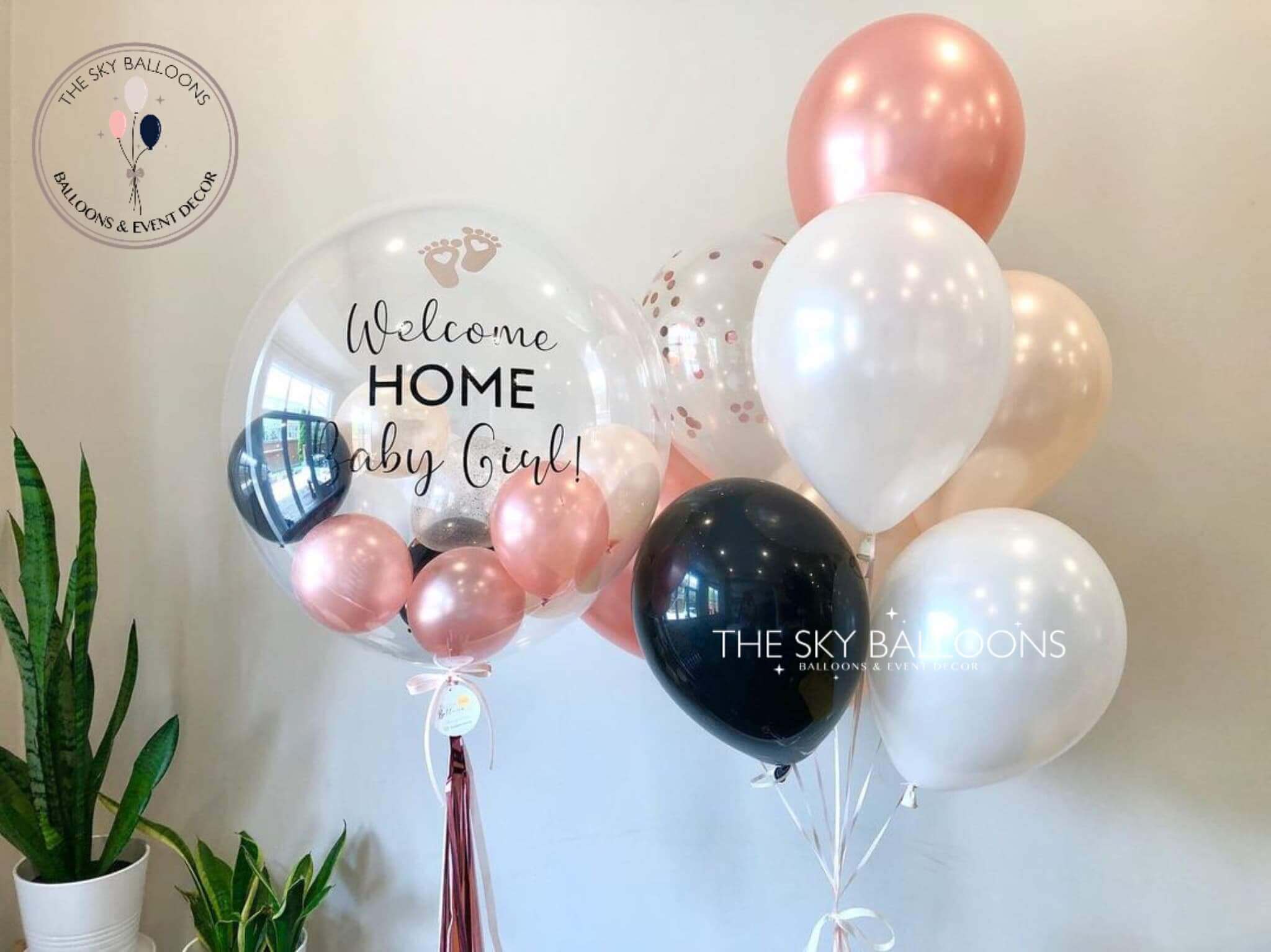A bouquet of latex balloons in various shades of pink, white, and black, filled with helium gas, with customized text that reads 'Welcome Home Baby Girl'. The balloons are arranged in a bunch with a pink ribbon and pearls, and the brand 'The Sky Balloons' is visible.