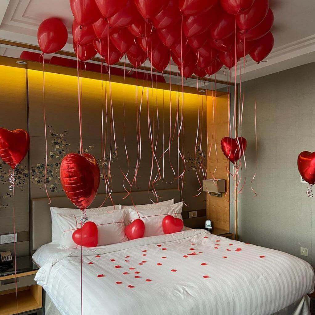 A hotel room decorated with red ceiling balloons, heart-shaped foil balloons, and scattered heart-shaped decorations on the floor, with the bed in the center of the room.