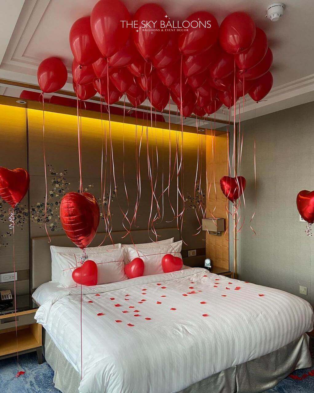 A hotel room decorated with red ceiling balloons, heart-shaped foil balloons, and scattered heart-shaped decorations on the floor, with the bed in the center of the room.