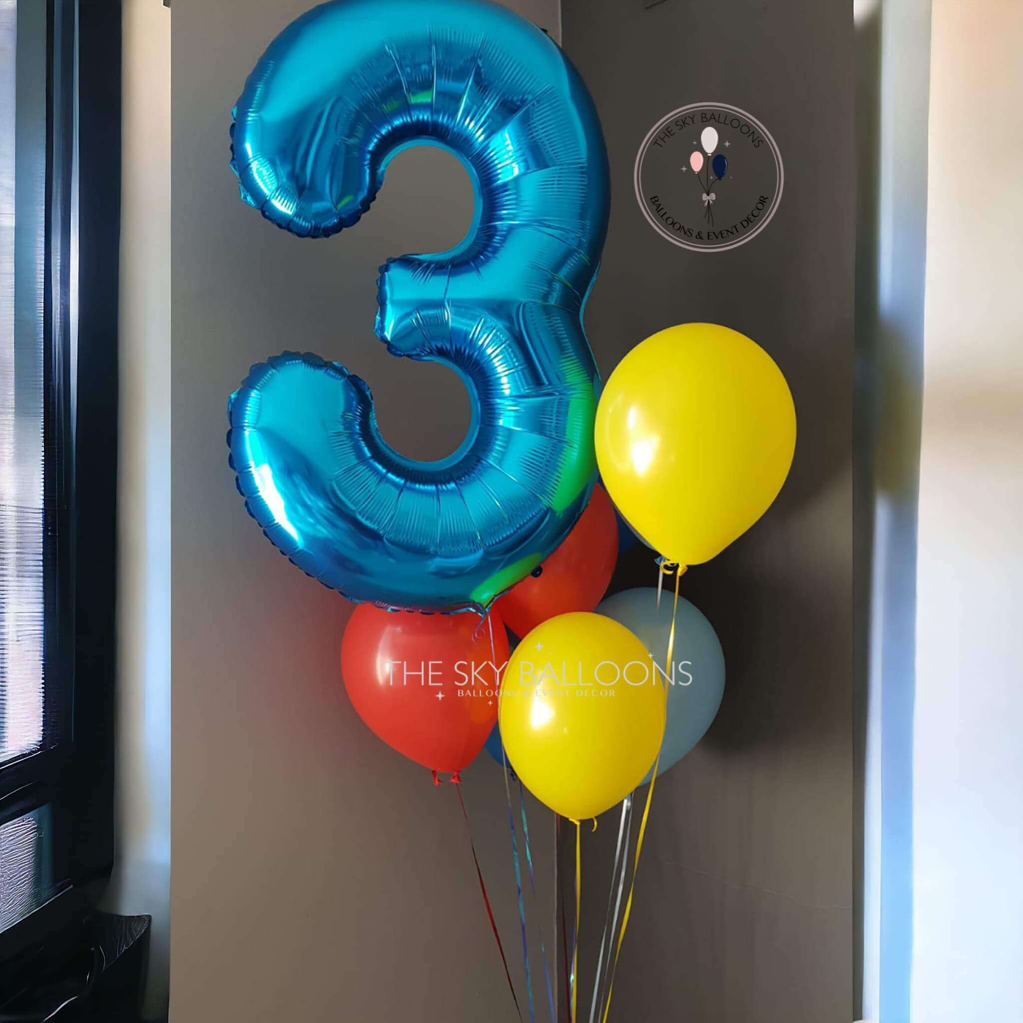 A balloon bouquet featuring a large blue number 3 balloon, with smaller yellow and red balloons tied to it, placed against a wall with a digital clock in the background.