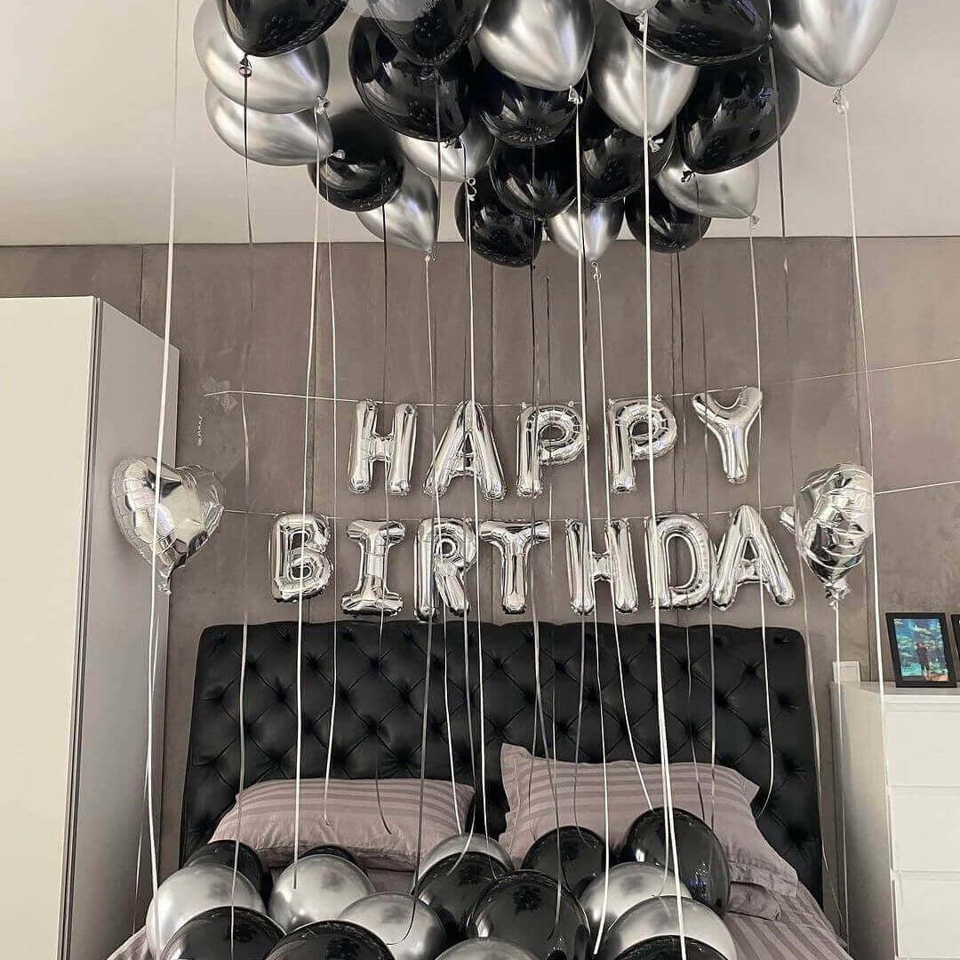A bedroom setup with silver and black balloons, including a 'HAPPY BIRTHDAY' banner, ceiling balloons, and heart-shaped foil balloons.