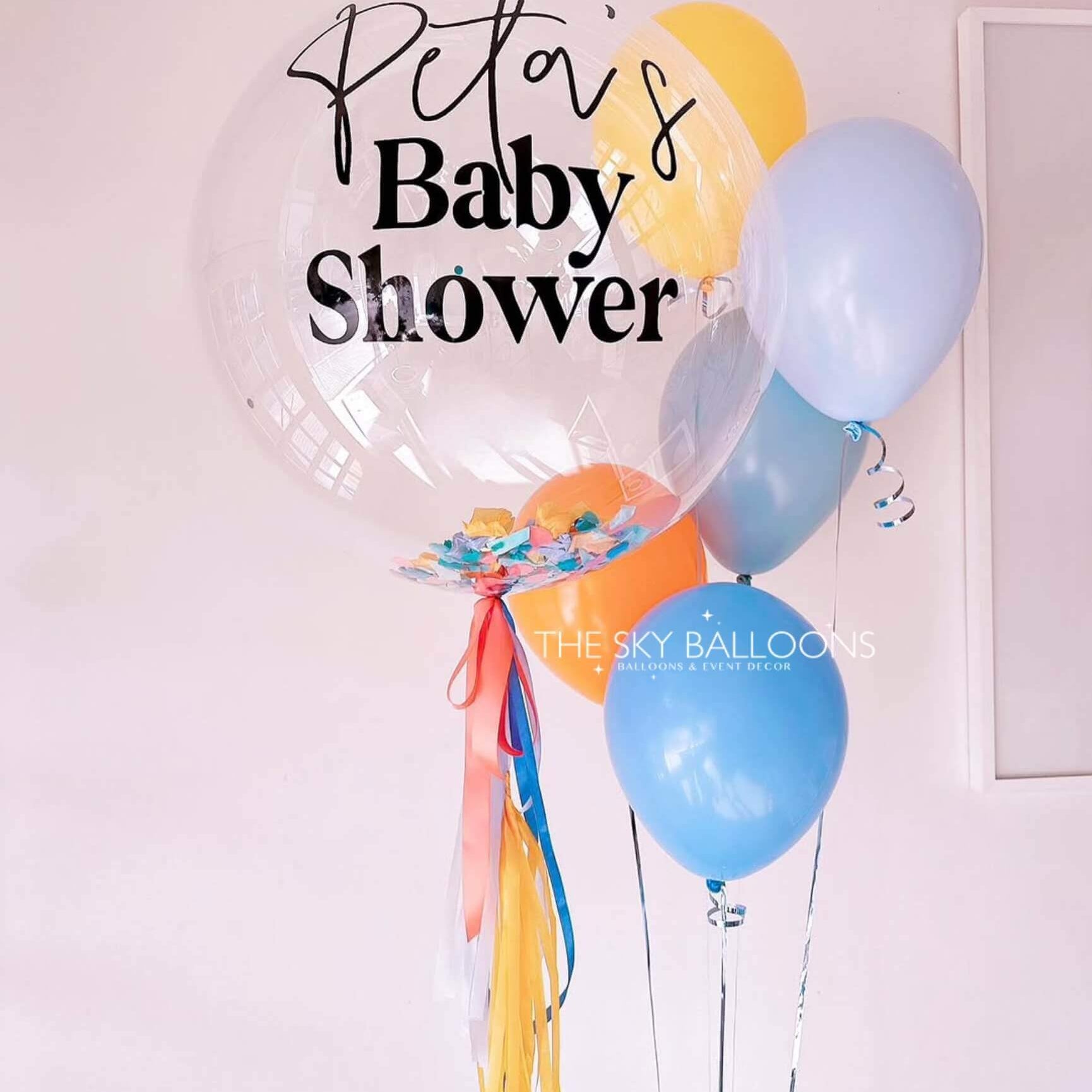 Clear balloon with 'Peta's Baby Shower' text and colorful balloons against a light pink wall.