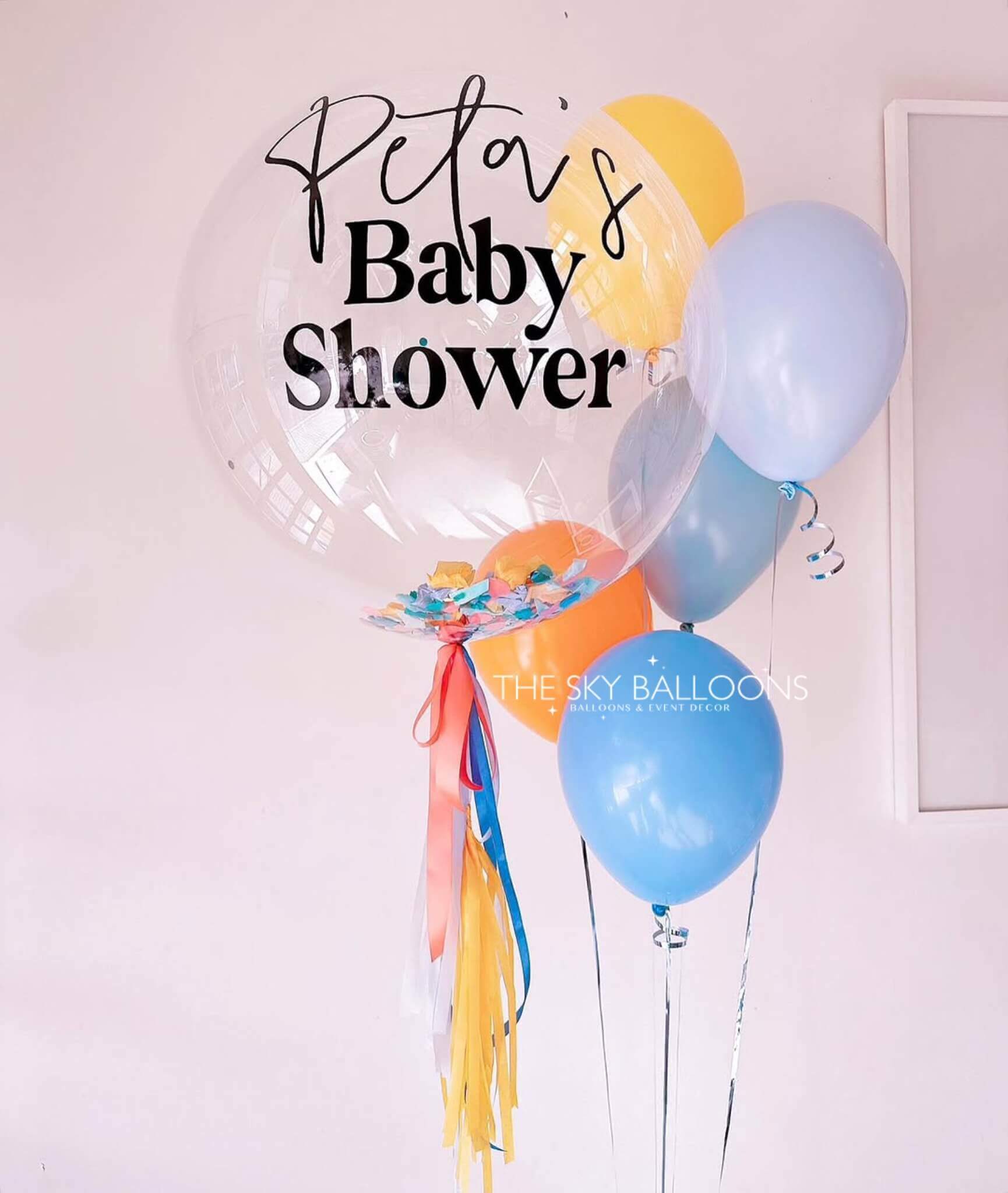 Clear balloon with 'Peta's Baby Shower' text and colorful balloons against a light pink wall.