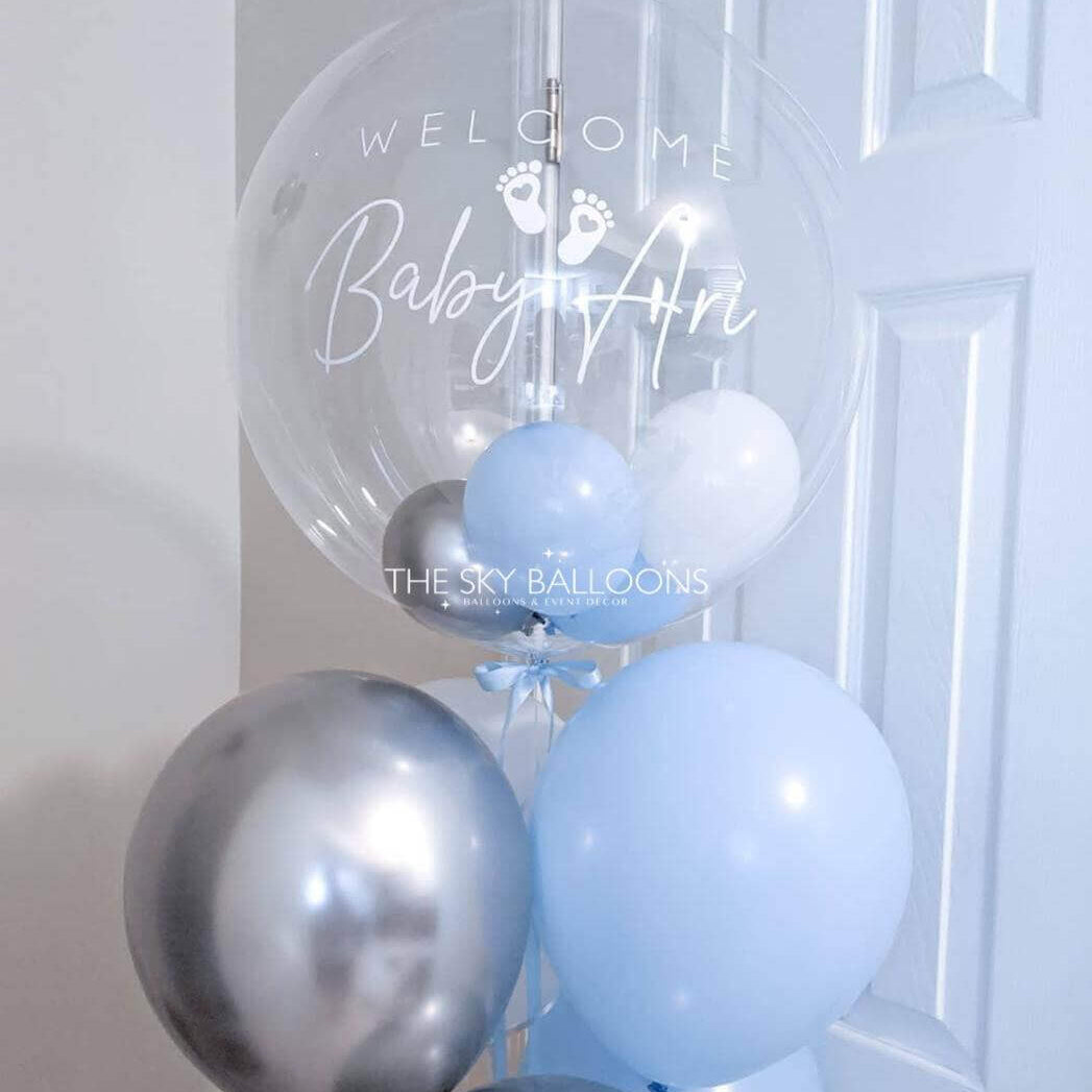 Clear balloon with 'Welcome Baby Ari' text and blue and silver balloons against a white door.