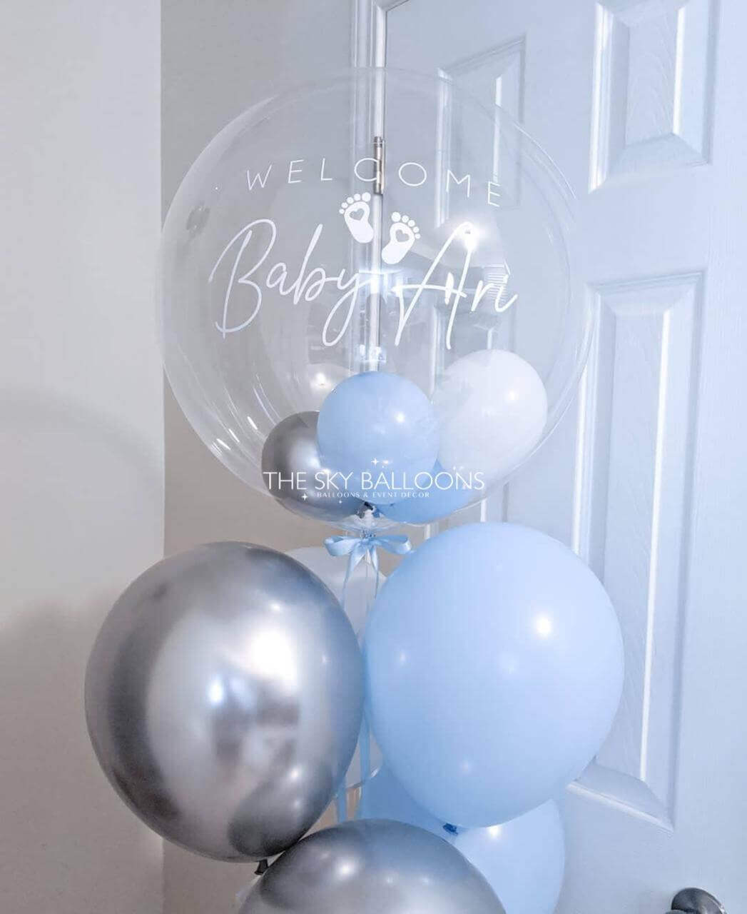 Clear balloon with 'Welcome Baby Ari' text and blue and silver balloons against a white door.