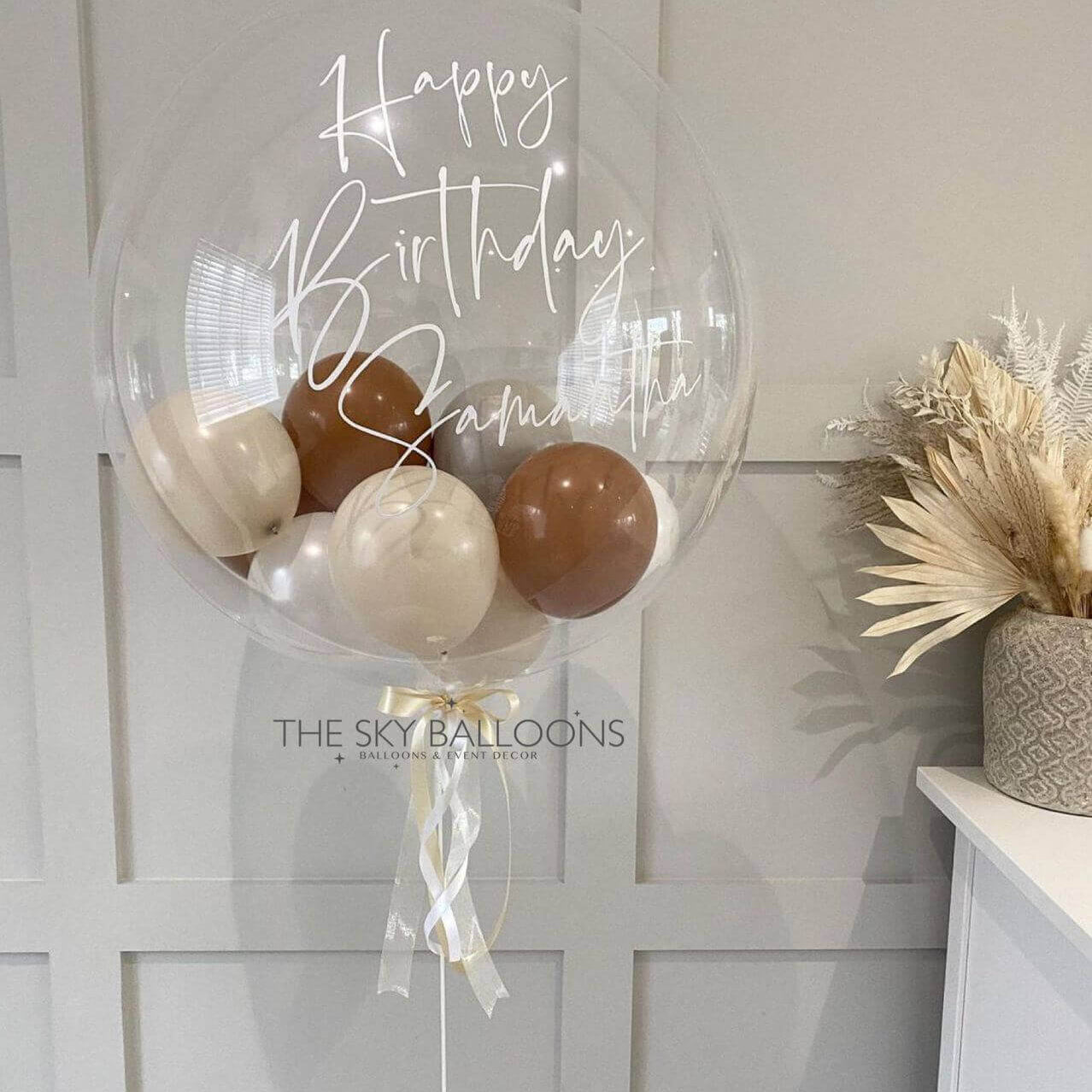 A large clear balloon with 'Happy Birthday' and a name written on it, containing smaller balloons in brown and white, displayed indoors.