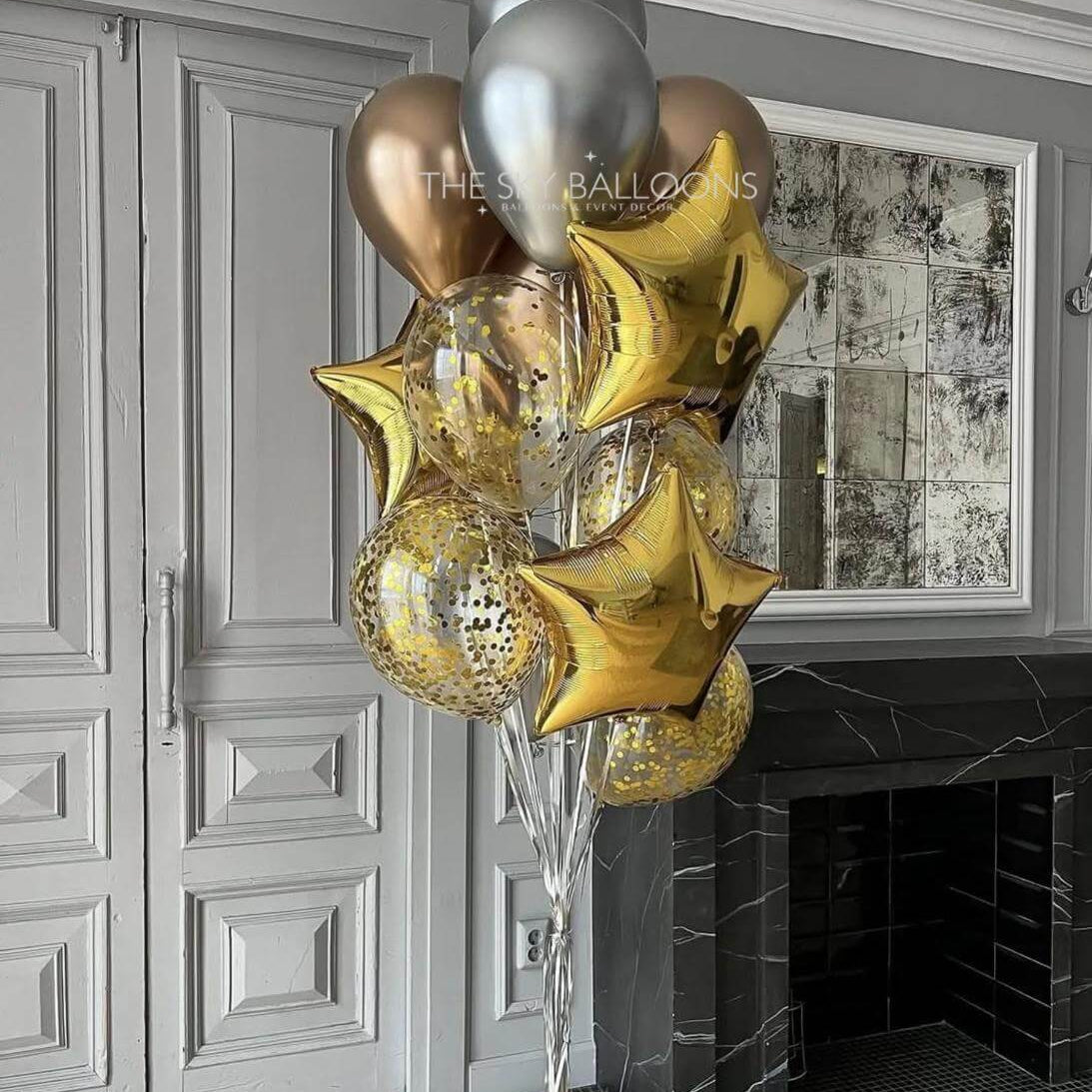 Decorative arrangement of gold, silver, and clear balloons in a room with a fireplace and framed picture on the wall.