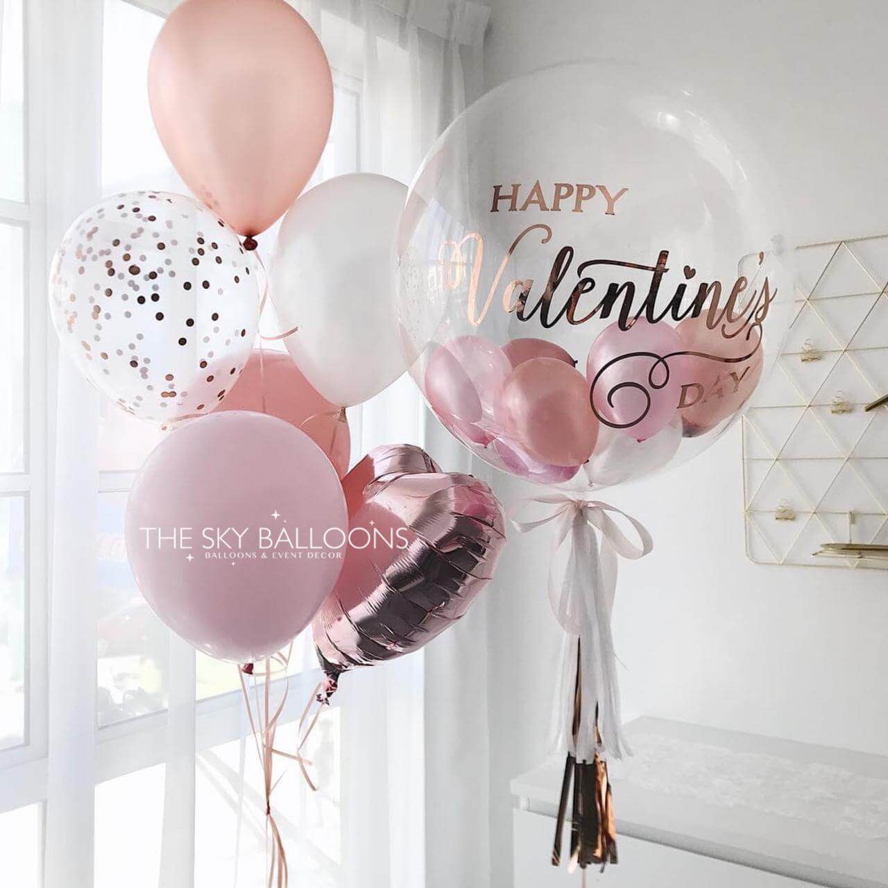 Decorative balloons with 'Happy Valentine's Day' text and confetti, tied together with ribbons against a light background.