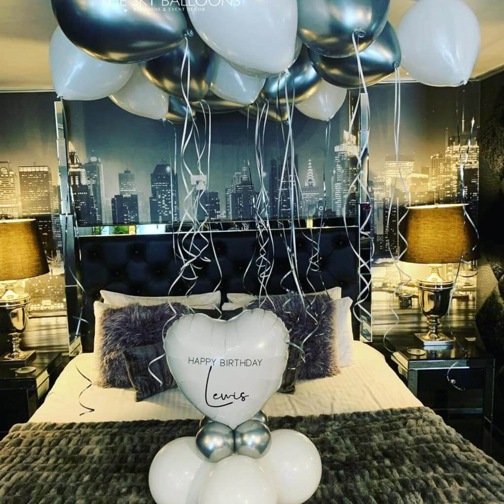Decorative setup with balloons and a heart-shaped balloon on a bed in a room with cityscape wallpaper.