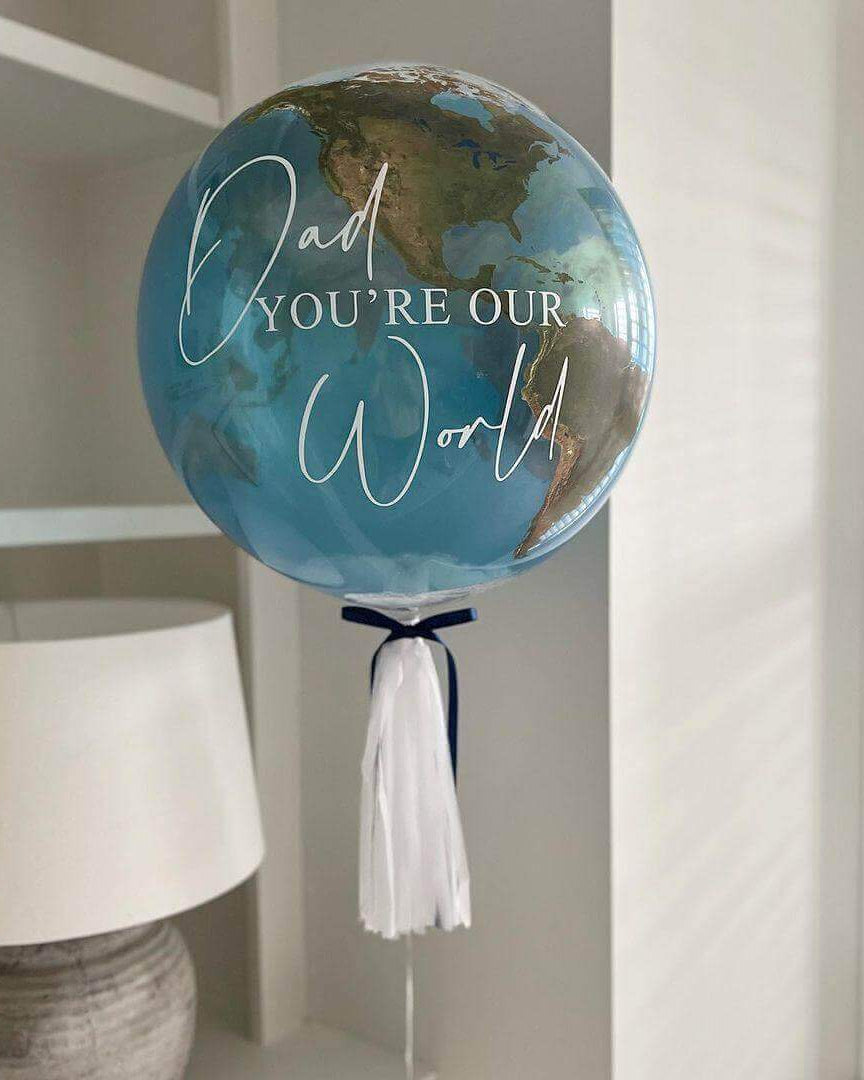 A balloon with a design resembling the Earth's globe and the text 'Dad you're our world' printed on it, adorned with tassels.