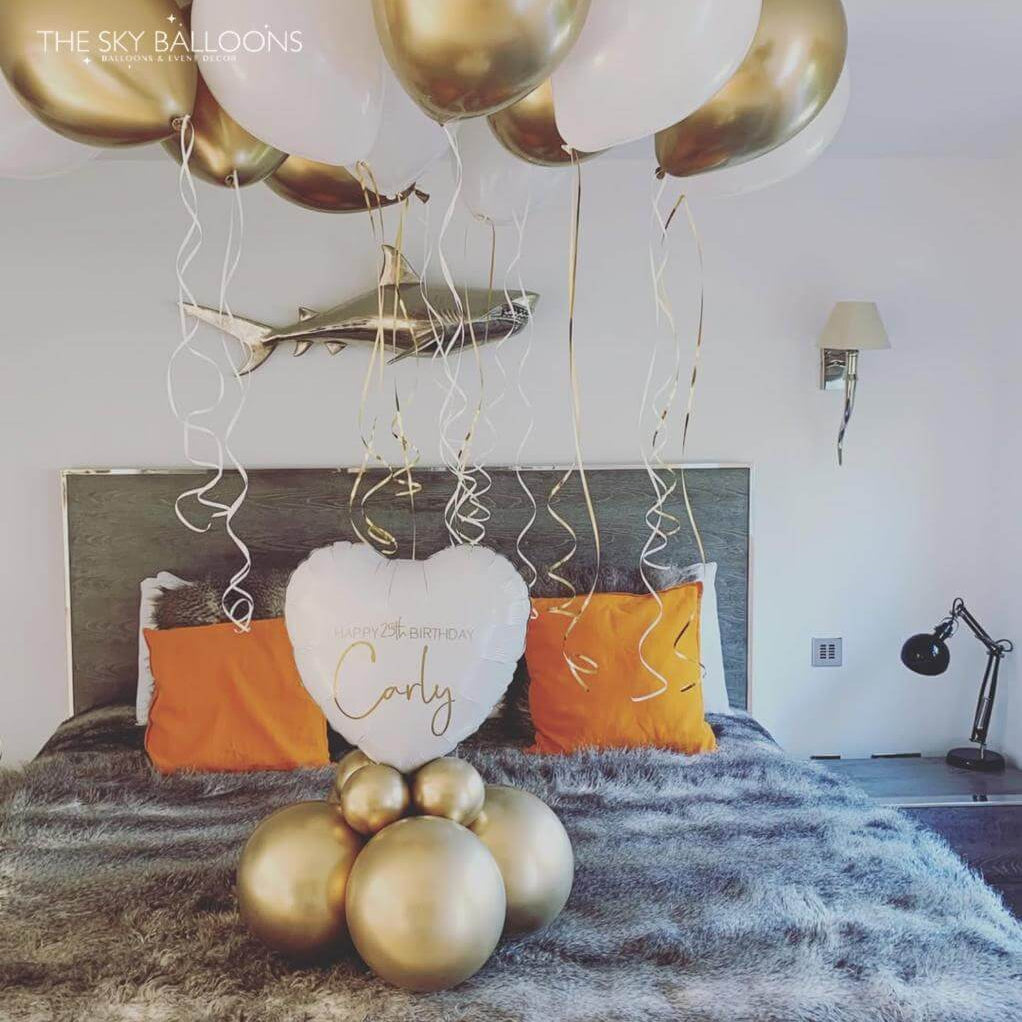 Decorative setup with gold and white balloons and a heart-shaped pillow on a bed.