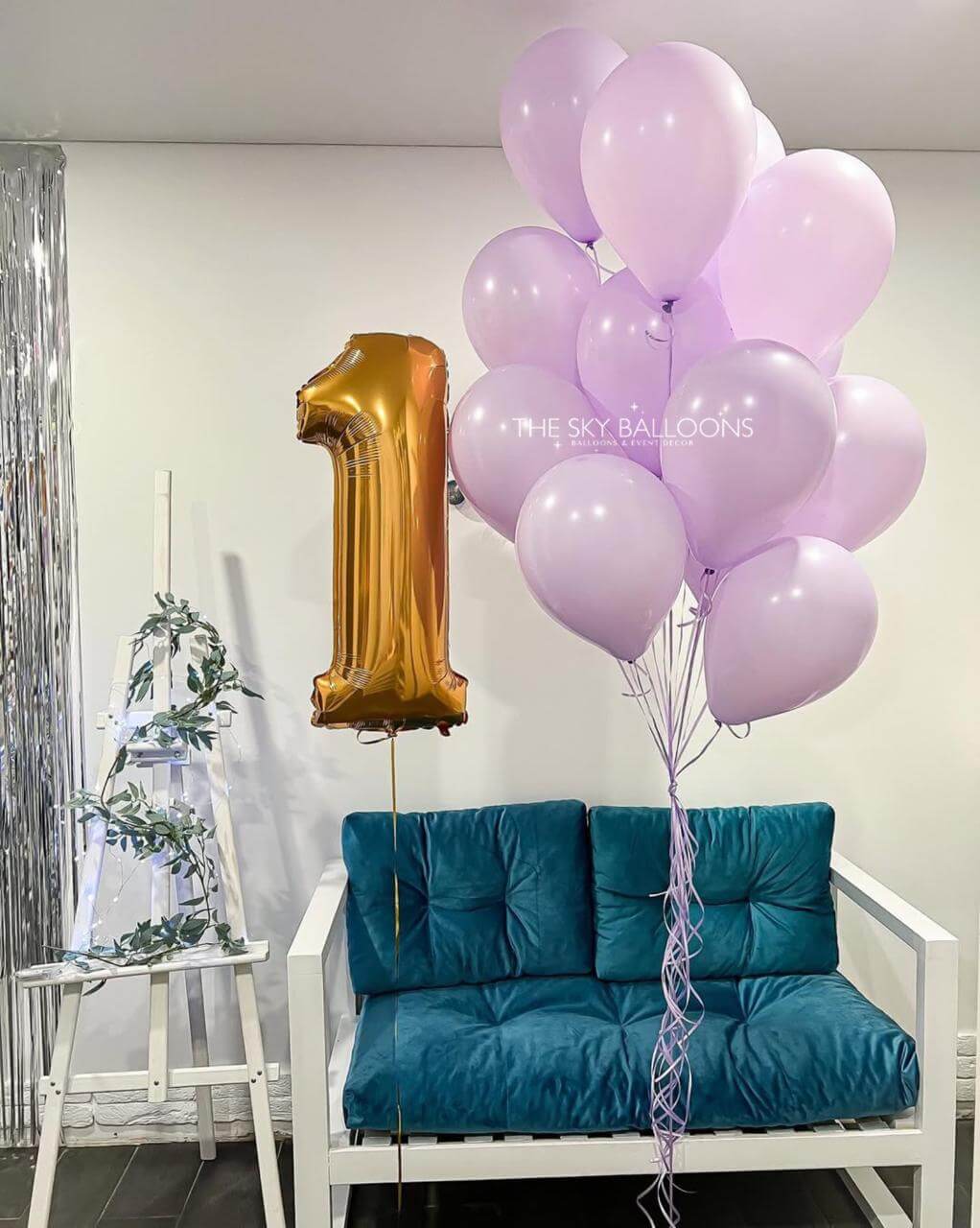 Gold number one balloon with pink balloons on a teal couch against a white wall.