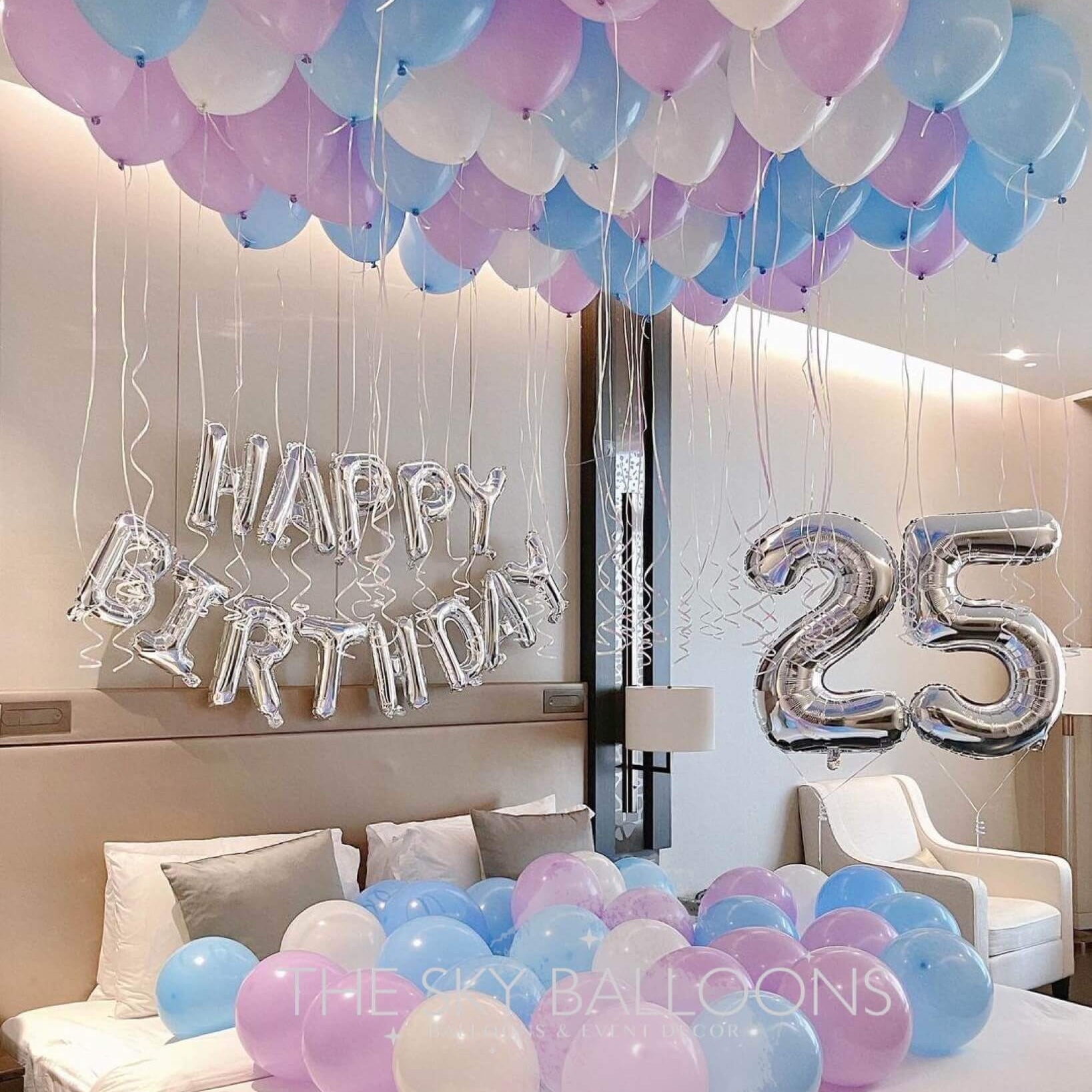 A room decorated with a 'Happy Birthday' balloon arrangement, featuring large numeral balloons '25', and a variety of colored balloons on the floor and ceiling.