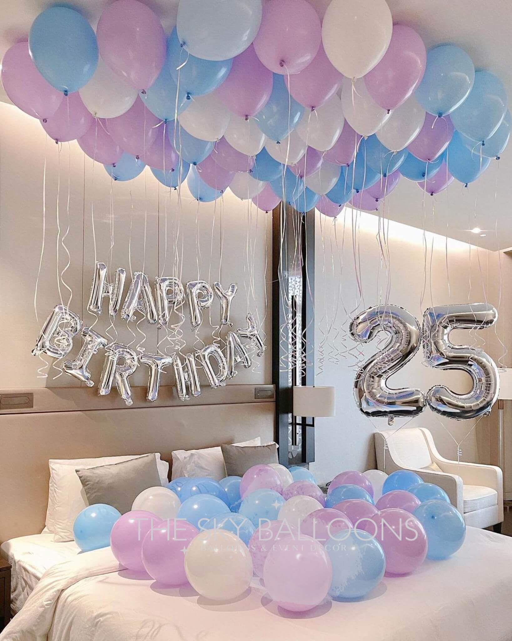 A room decorated with a 'Happy Birthday' balloon arrangement, featuring large numeral balloons '25', and a variety of colored balloons on the floor and ceiling.