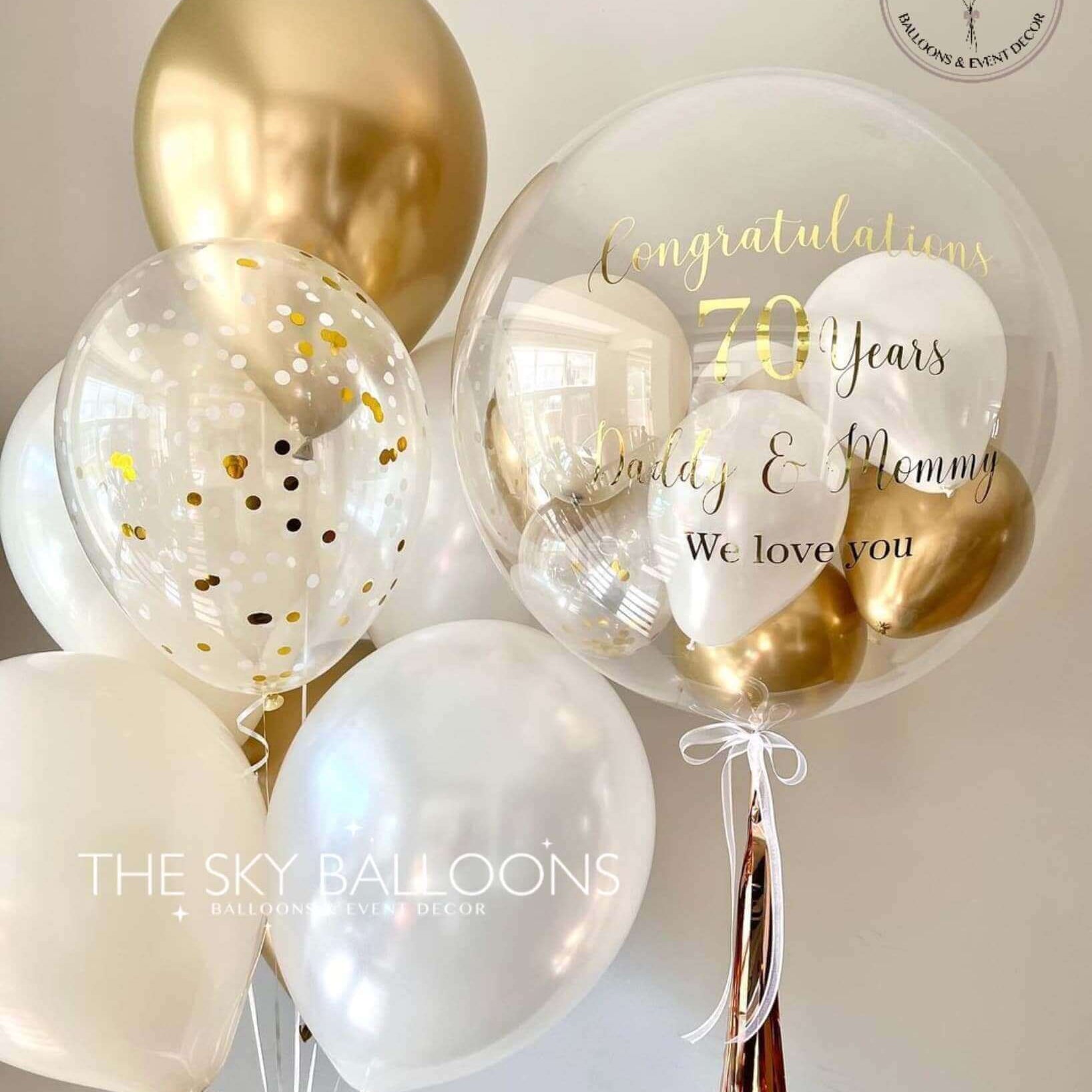 A bouquet of latex balloons filled with helium gas, featuring white and gold colors with printed text that reads 'Happy 10th Anniversary'. There are also smaller balloons and tassels visible within the larger balloons.