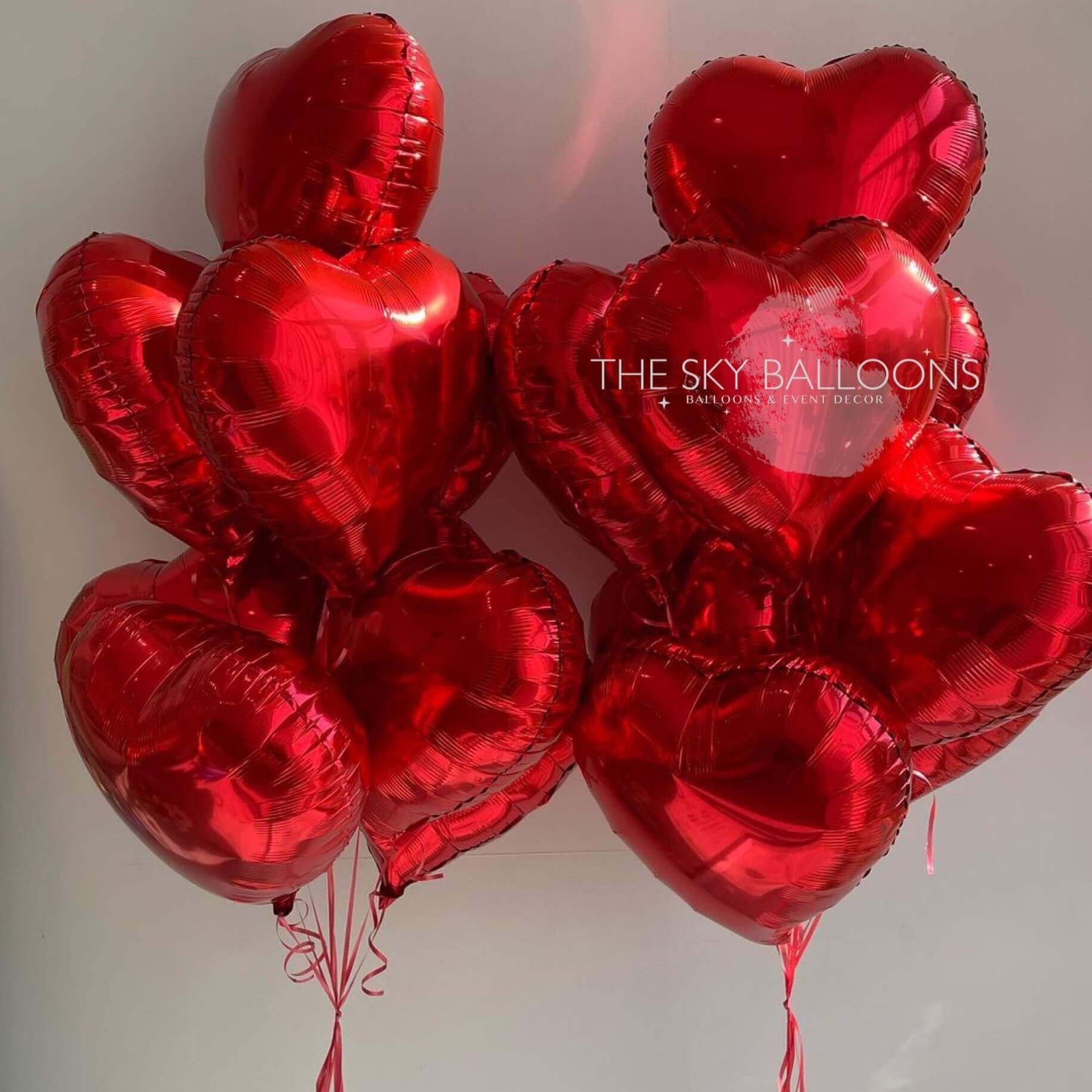 A bunch of inflated red heart-shaped foil balloons with the text 'THE SKY BALLOONS' on a reflective surface.