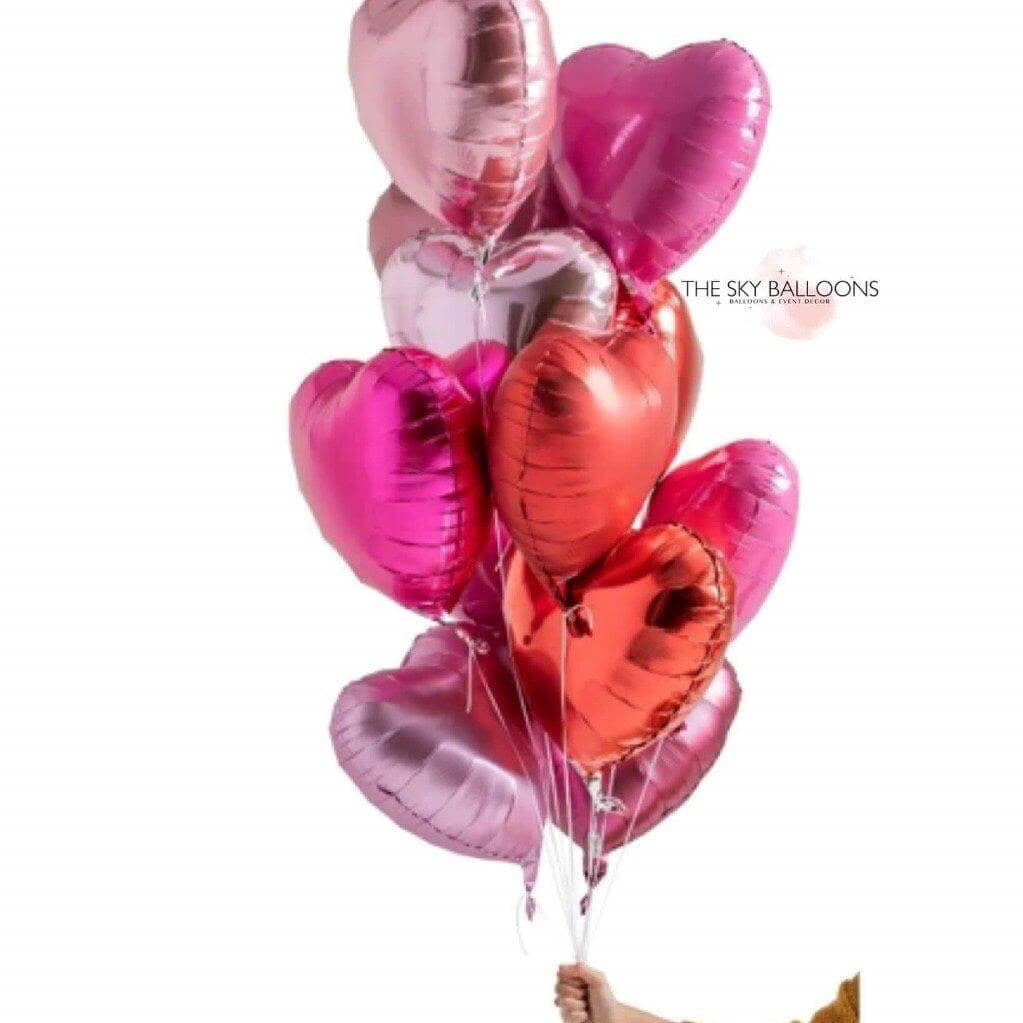 A bouquet of heart-shaped balloons in various shades of pink, silver, and gold, held by a hand against a white background.