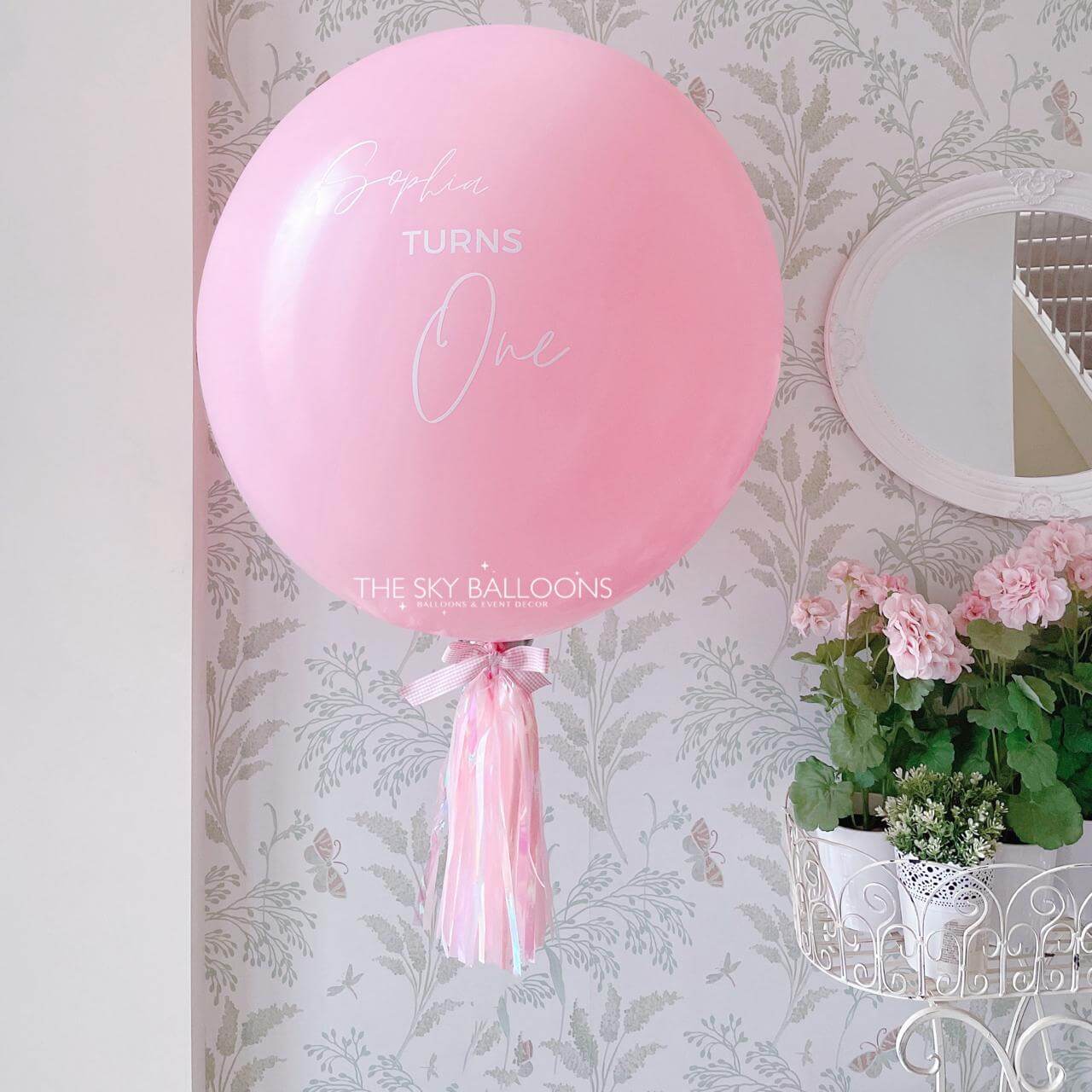 Pink balloon with 'Happy Turns One' text against a floral wallpaper background