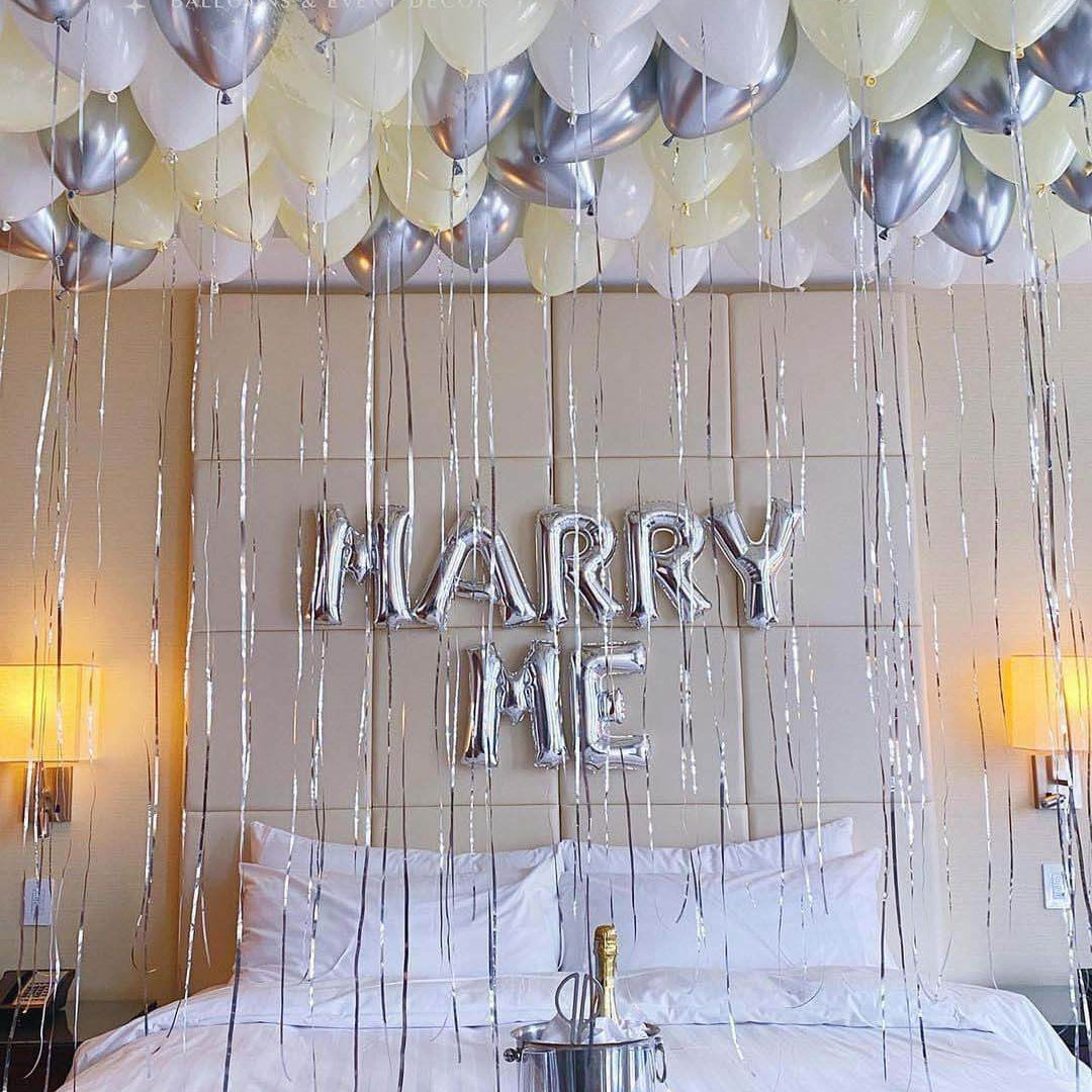 A bedroom setup with silver, chrome, ivory, and white balloons forming a 'MARRY ME' banner above the bed.