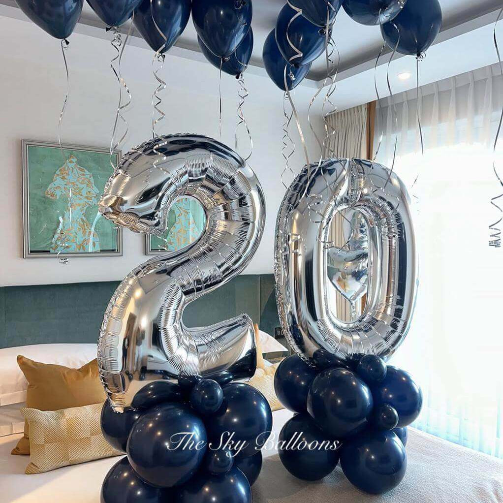 Elegant 20th birthday balloon decoration featuring large silver foil number '20' balloons on navy blue balloon bases, with matching navy blue helium balloons floating on the ceiling with silver ribbons, displayed in a modern living room setting