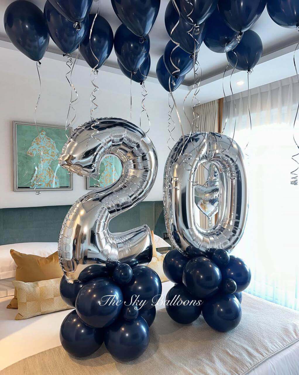Elegant 20th birthday balloon decoration featuring large silver foil number '20' balloons on navy blue balloon bases, with matching navy blue helium balloons floating on the ceiling with silver ribbons, displayed in a modern living room setting