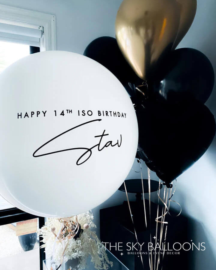 A bouquet of balloons featuring black and gold colored latex balloons with one personalized balloon that has text on it.