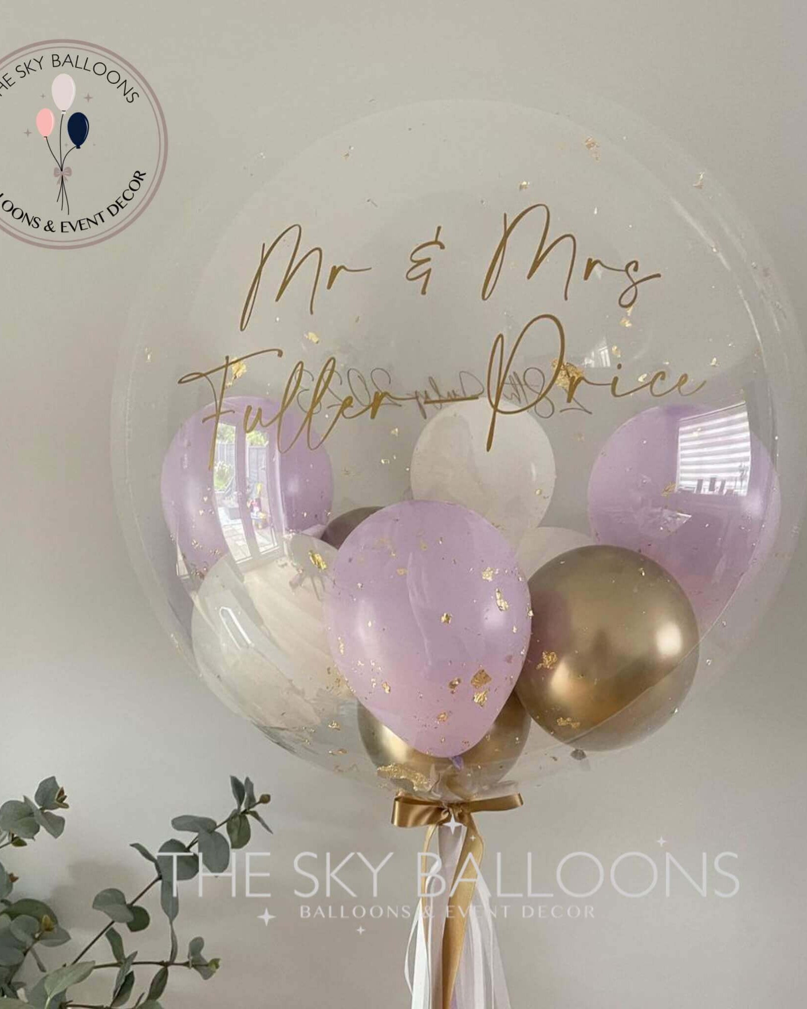 A customized pastel lilac bubble balloon with gold and purple tassels, filled with helium, alongside smaller balloons. There is a golden balloon bouquet with the text 'Mr & Mrs Fullerton Price' written on it.