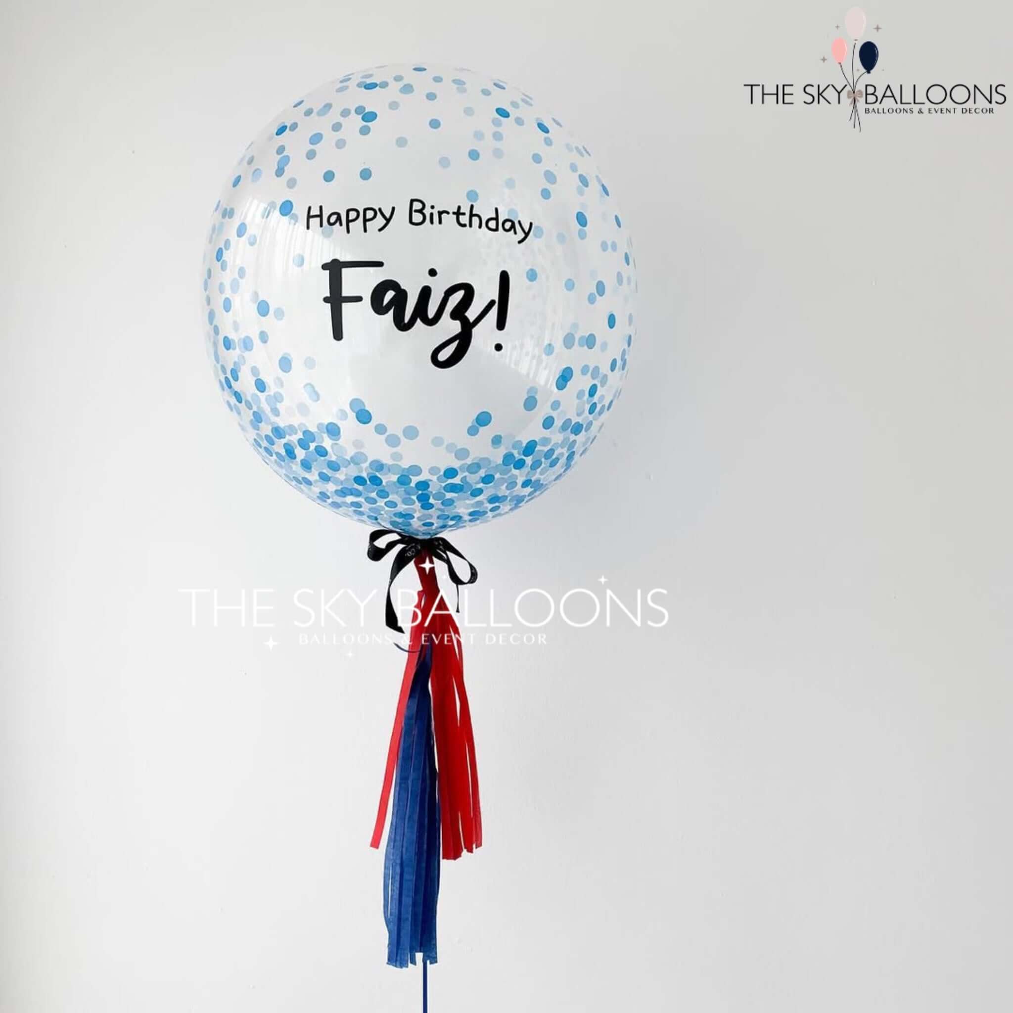A personalized round balloon with a blue dotted pattern, black text reading 'Happy Birthday', and a name 'Fay'. The balloon is attached to a red and blue tassel.