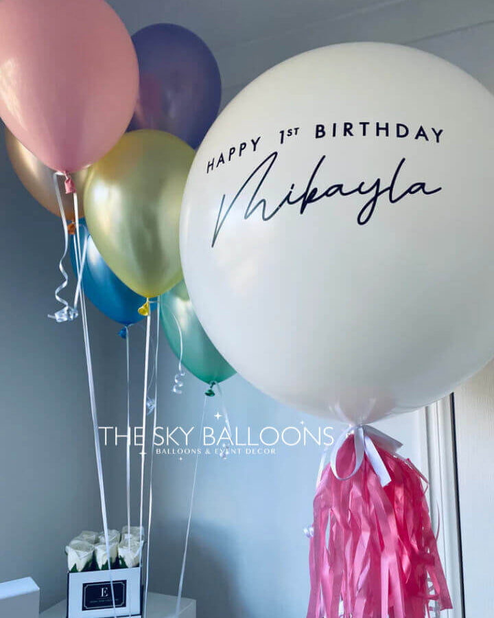 A set of balloons in various colors including pink, gold, blue, and white, with one large personalized balloon reading 'Happy 1st Birthday Mikayla', and attached pink weight tassels.