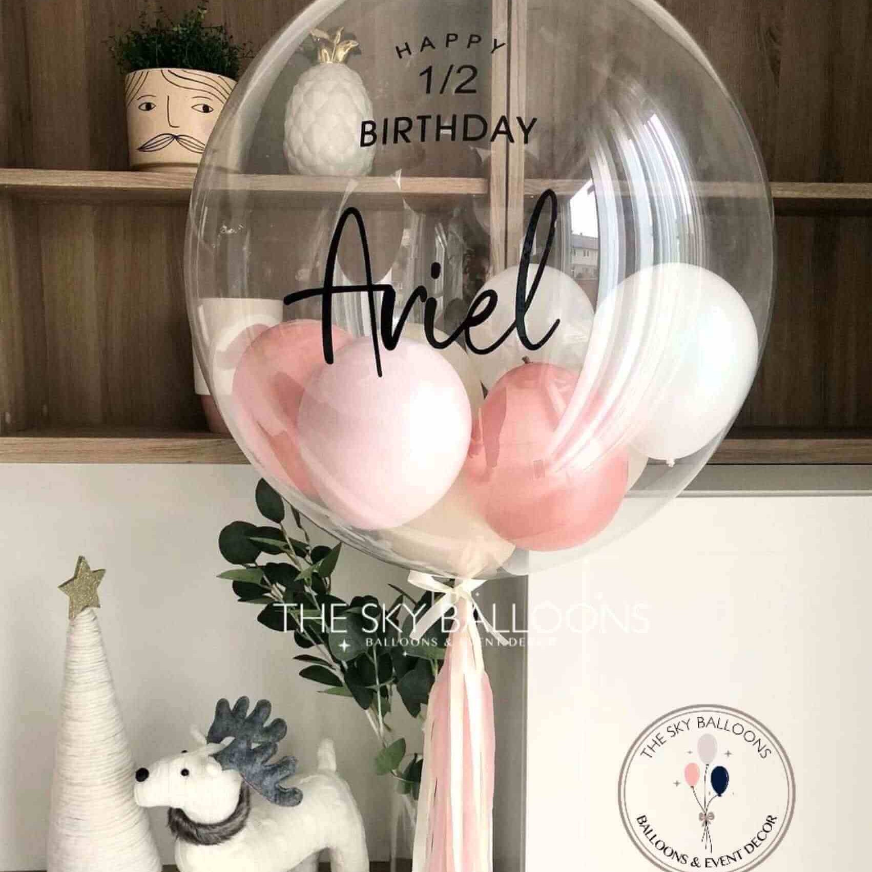 A rose gold bubble balloon with the name 'Aniel' printed on it, surrounded by smaller pink balloons, placed near a decorative tree and a white balloon, with a 'Happy Birthday' sign in the background.