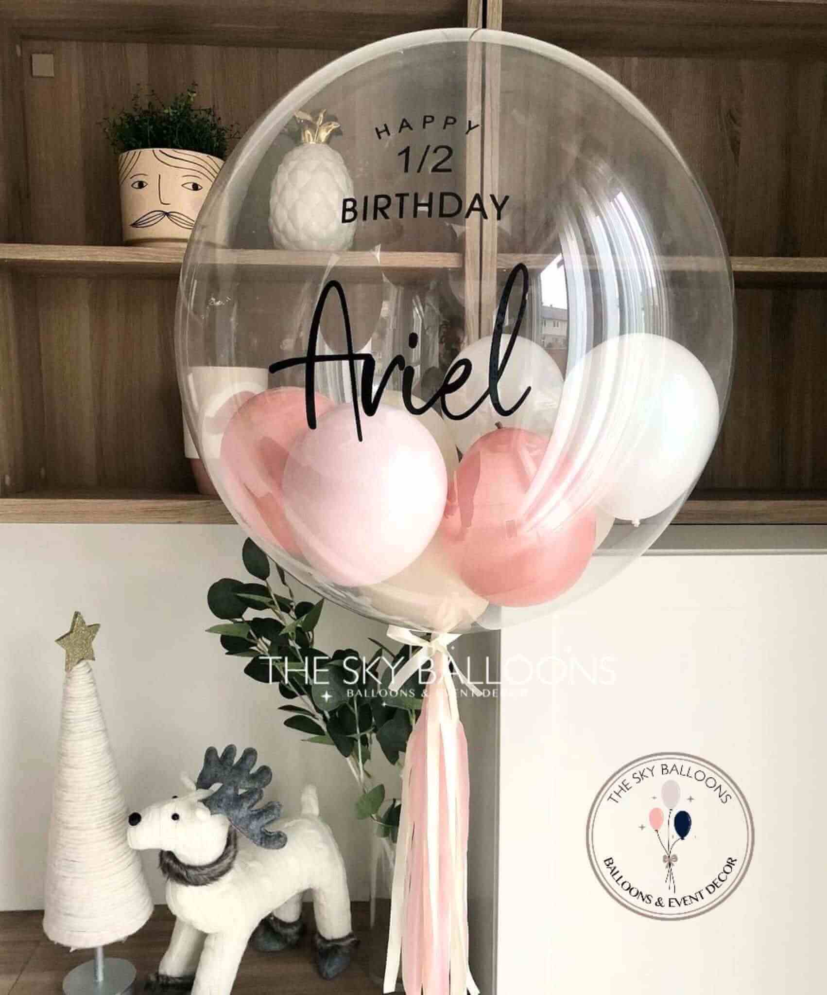 A rose gold bubble balloon with the name 'Aniel' printed on it, surrounded by smaller pink balloons, placed near a decorative tree and a white balloon, with a 'Happy Birthday' sign in the background.