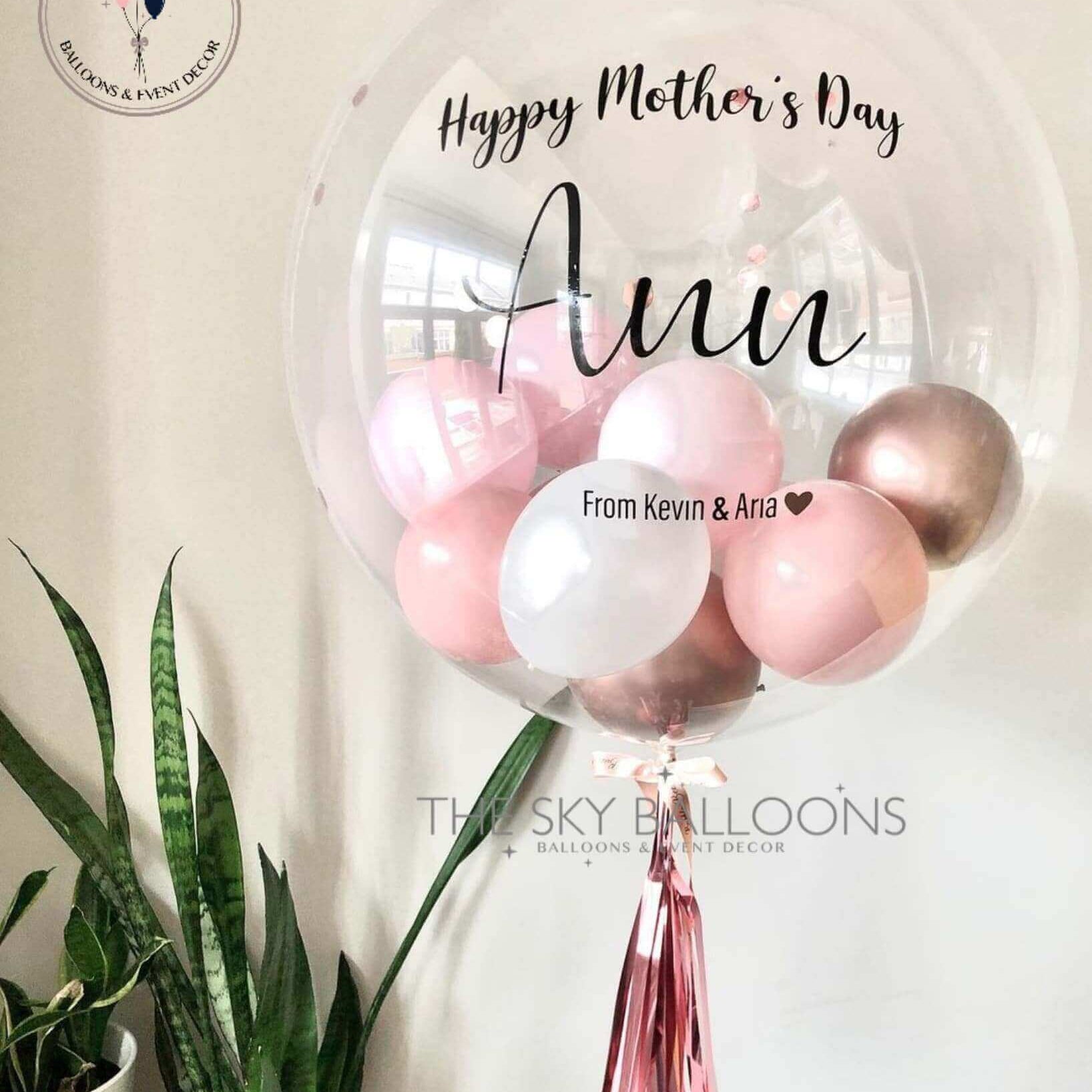 A customized balloon with 'Happy Mother's Day' text, alongside smaller balloons and tassels, all in shades of pink and gold, with a plant decor in the background.
