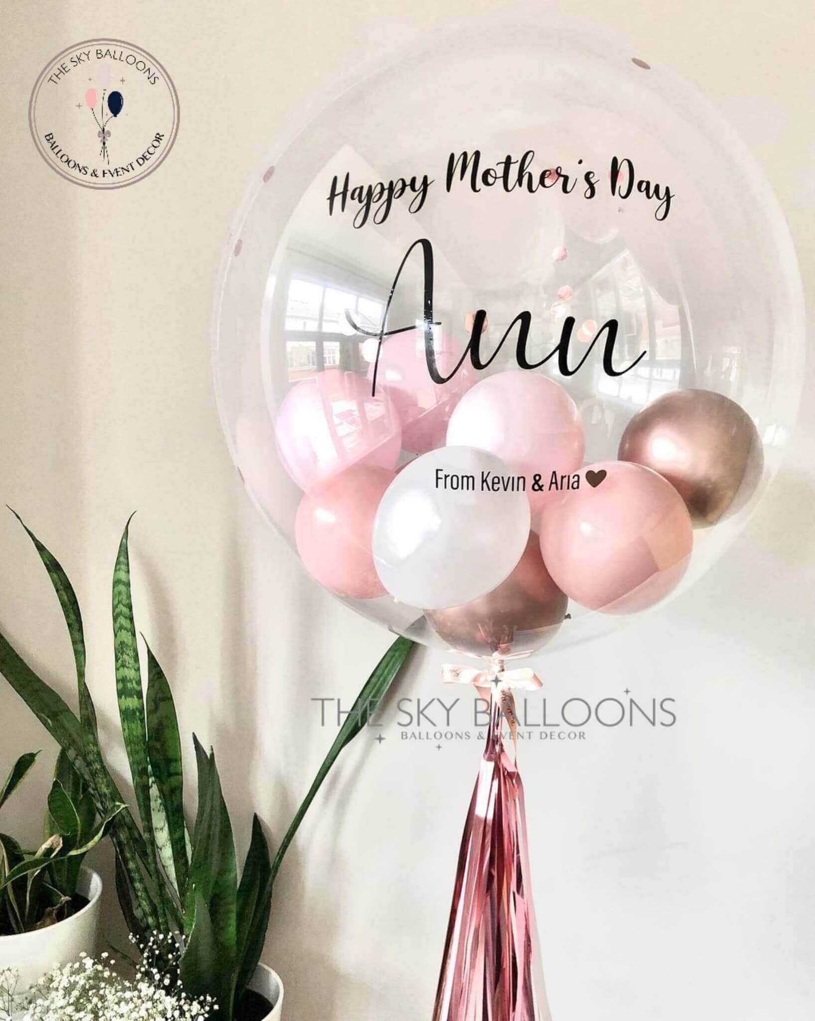 A customized balloon with 'Happy Mother's Day' text, alongside smaller balloons and tassels, all in shades of pink and gold, with a plant decor in the background.