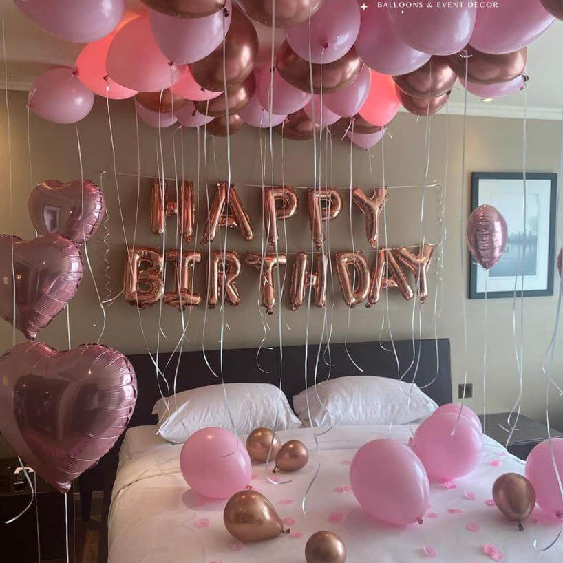A room decorated with pink and gold balloons, including a 'Happy Birthday' banner, ceiling balloons, and heart-shaped balloons.