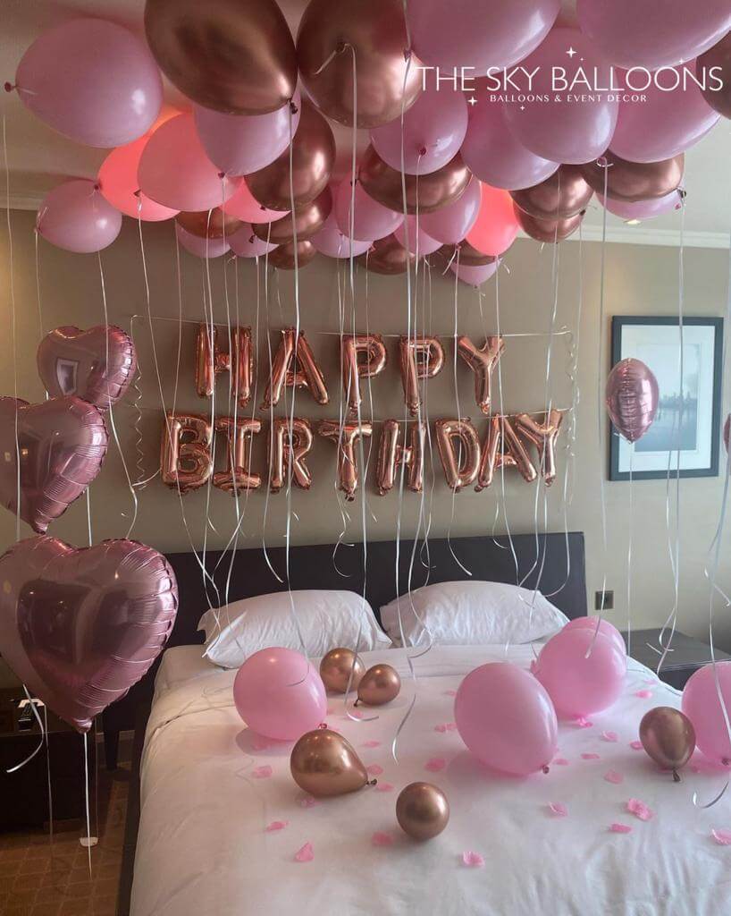 A room decorated with pink and gold balloons, including a 'Happy Birthday' banner, ceiling balloons, and heart-shaped balloons.