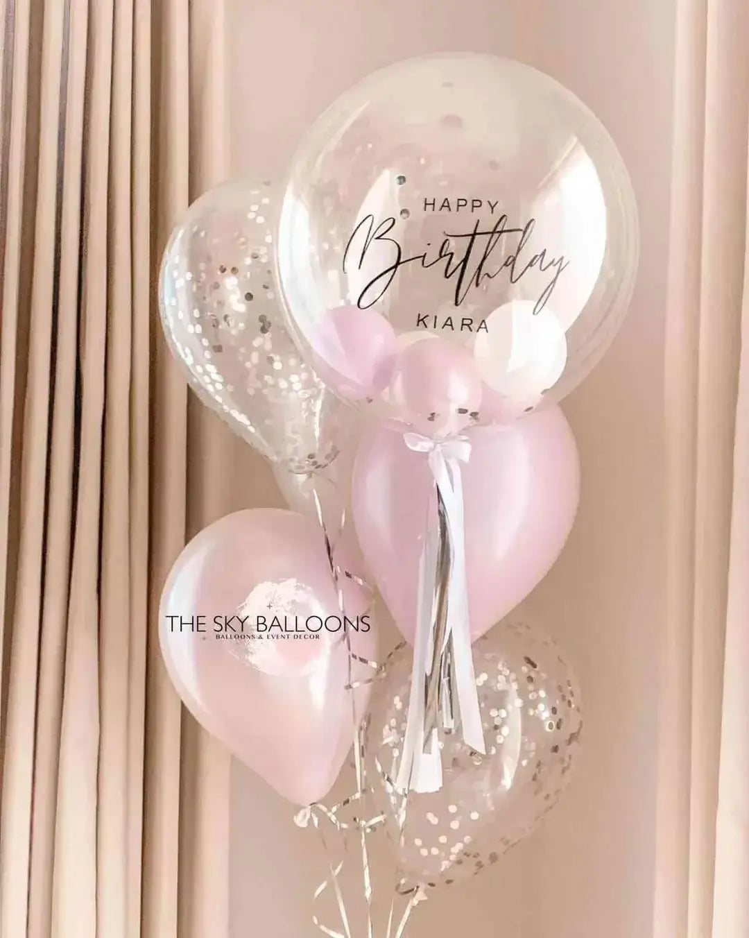 A balloon bouquet featuring a large pink customized balloon with the text 'Happy Birthday', two smaller clear balloons with pink confetti, and decorative tassels.