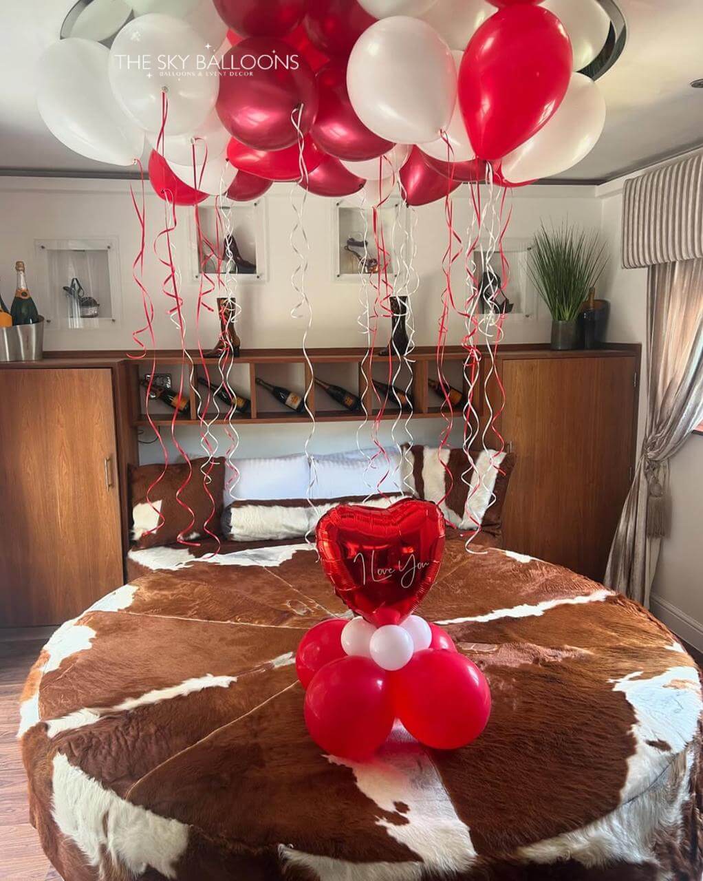 Red and white balloons with a heart-shaped balloon on a cowhide rug in a room.
