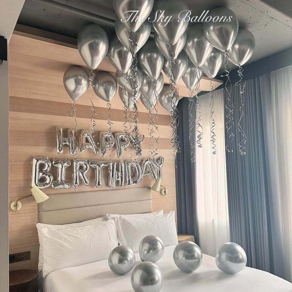 Sophisticated silver birthday balloon decoration featuring 'HAPPY BIRTHDAY' letter balloons with matching silver helium balloons floating on ceiling and chrome balloons arranged on white hotel bed in modern bedroom with wood accent wal