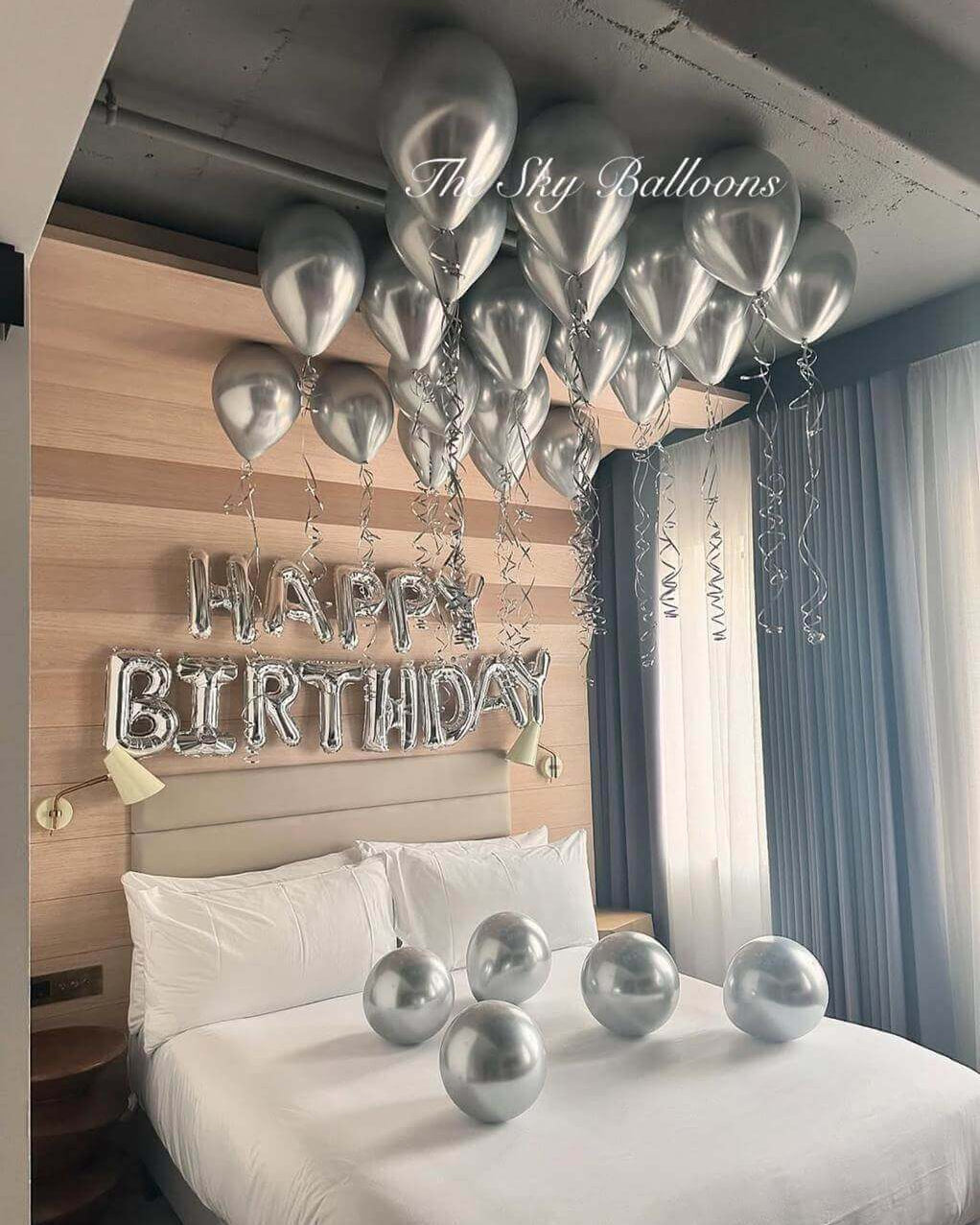 Sophisticated silver birthday balloon decoration featuring 'HAPPY BIRTHDAY' letter balloons with matching silver helium balloons floating on ceiling and chrome balloons arranged on white hotel bed in modern bedroom with wood accent wal
