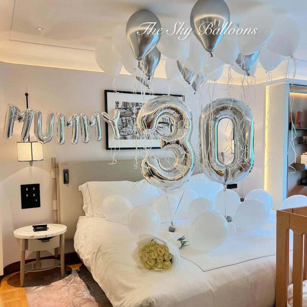 Elegant 30th birthday balloon decoration featuring silver 'MUM' letter balloons and large silver foil number '30' balloons with white and silver helium balloons floating on ceiling, displayed in modern hotel bedroom with white roses