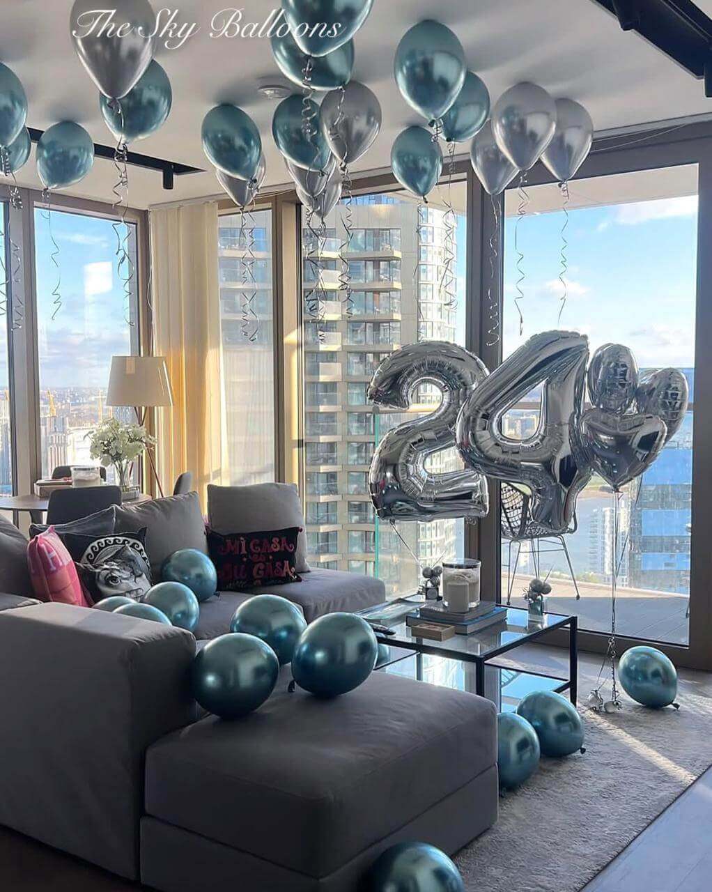 Decorative balloons, including large silver '24', in a living room with cityscape view.