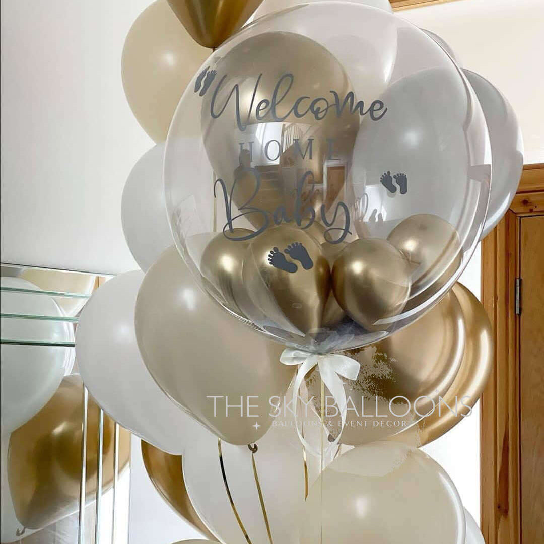 A bouquet of latex balloons in shades of gold, white, and transparent, with 'Welcome Home' text printed on a large gold balloon, tied with ribbons and helium-filled.