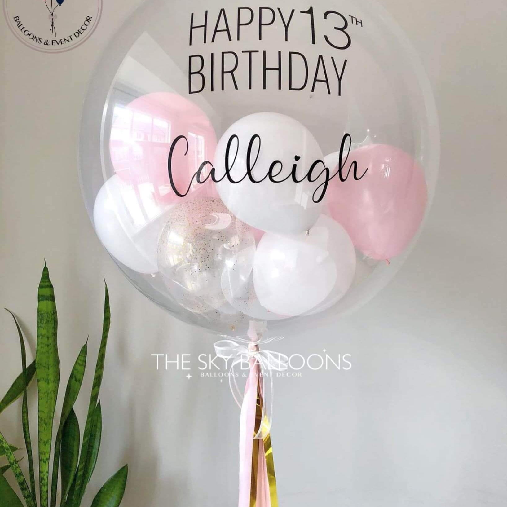 A customized round birthday balloon with 'Happy 13th Birthday' written on it, filled with helium and adorned with small pink and white balloons and tassels, placed against a neutral background.