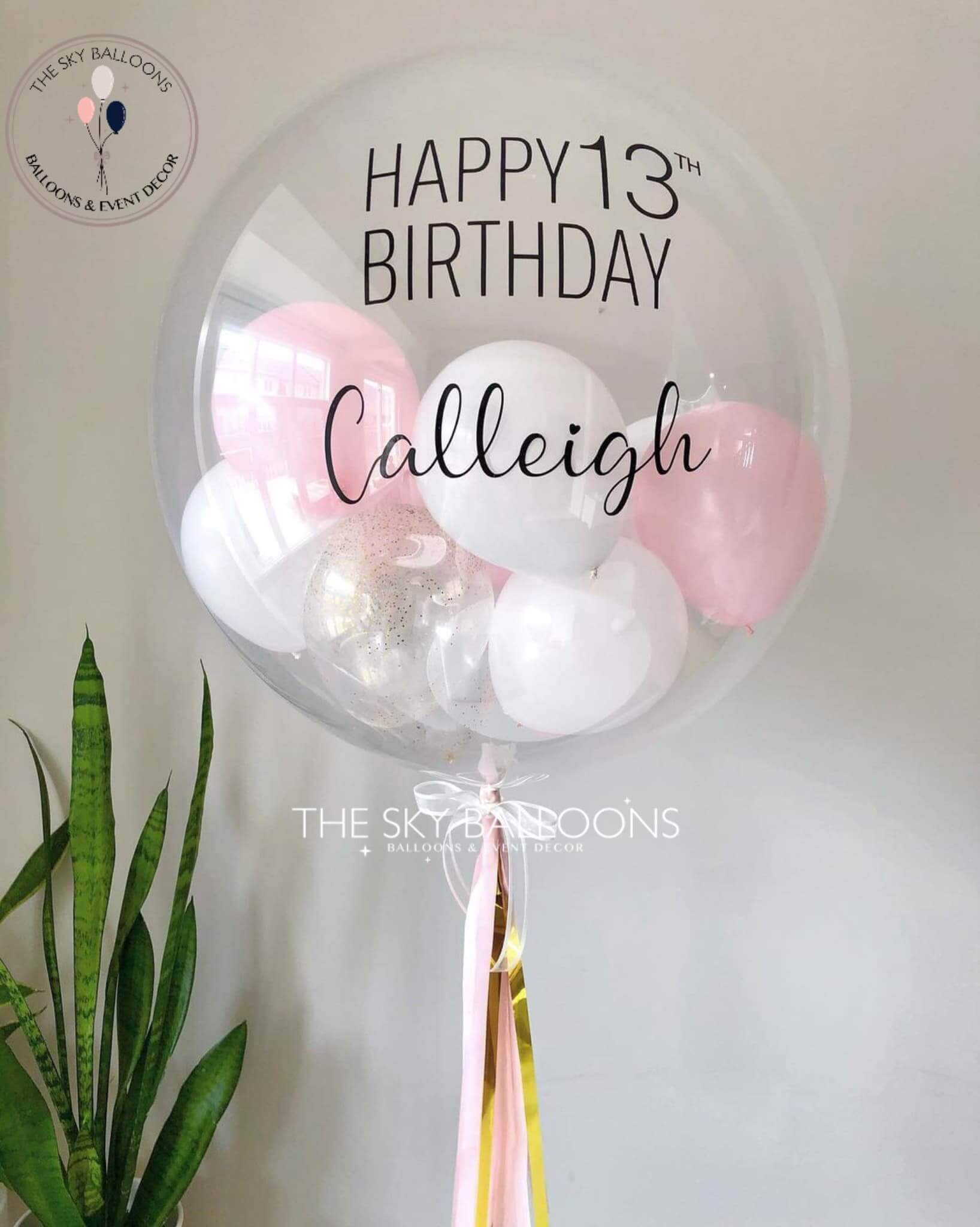 A customized round birthday balloon with 'Happy 13th Birthday' written on it, filled with helium and adorned with small pink and white balloons and tassels, placed against a neutral background.