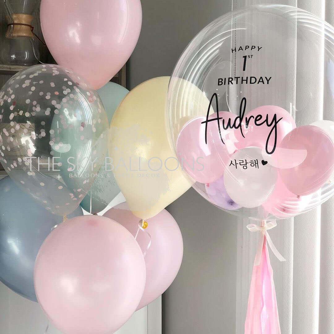 A bouquet of personalized balloons in shades of pink, blue, and transparent, with 'Happy Birthday' and a name 'Audrey' printed on a pink balloon, along with other balloons with different messages.